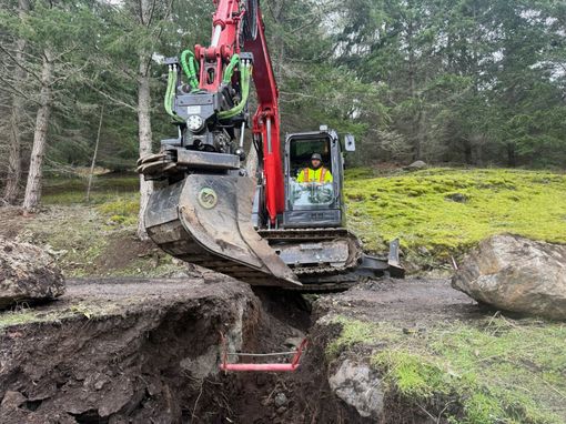 A yellow excavator using a specialized clamp attachment to lift and position a long black pipe on a gravel site.
