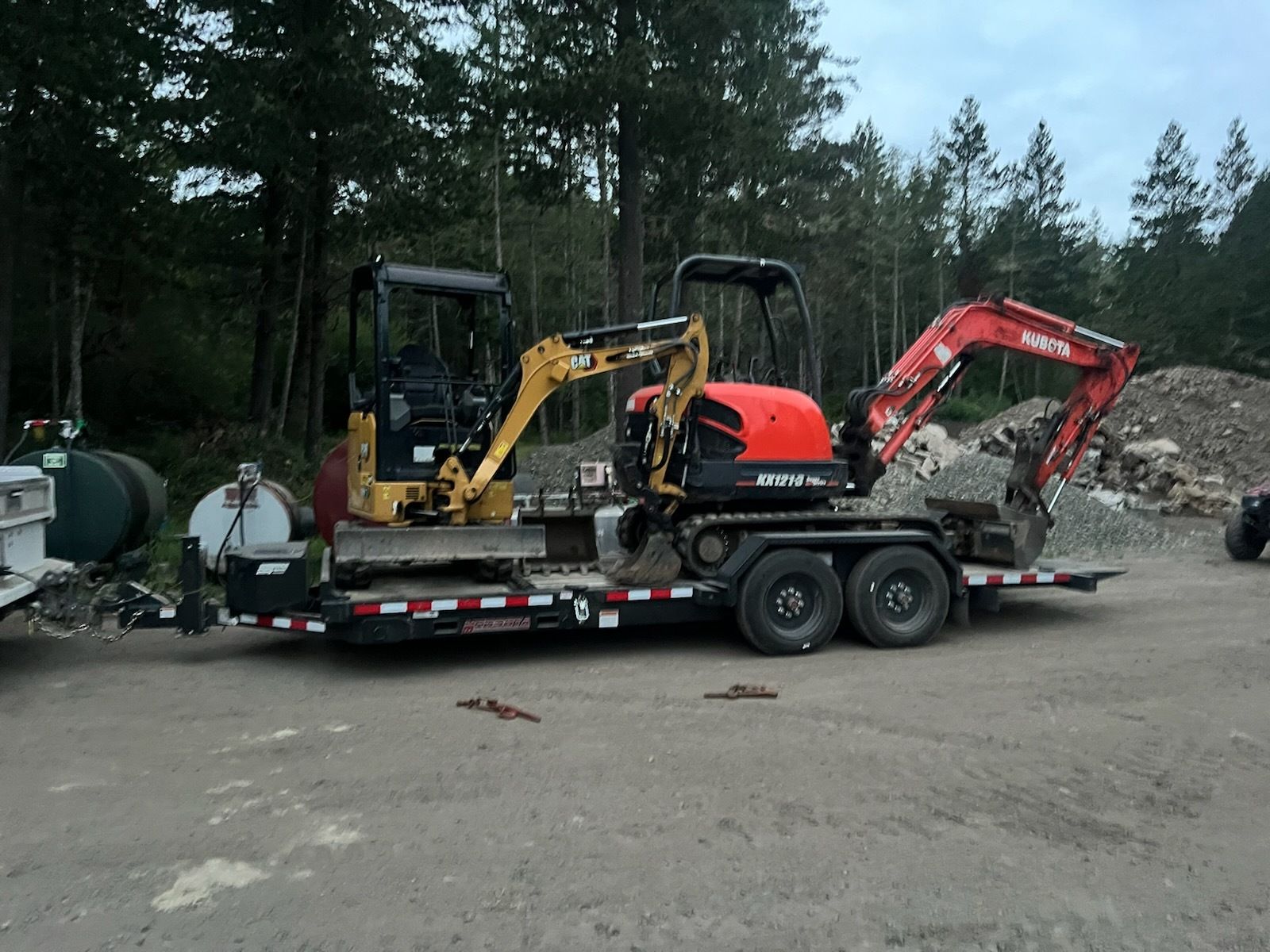 A flatbed trailer parked on a dirt lot carries a yellow excavator and a red excavator, with trees in the background.