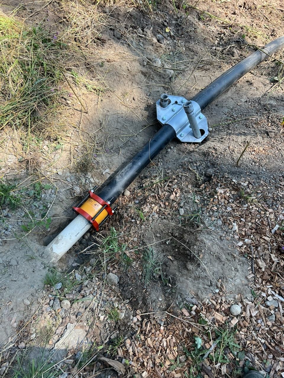 A metal pipe with a black-and-orange coupling and a mounting bracket lying on dirt and grass.