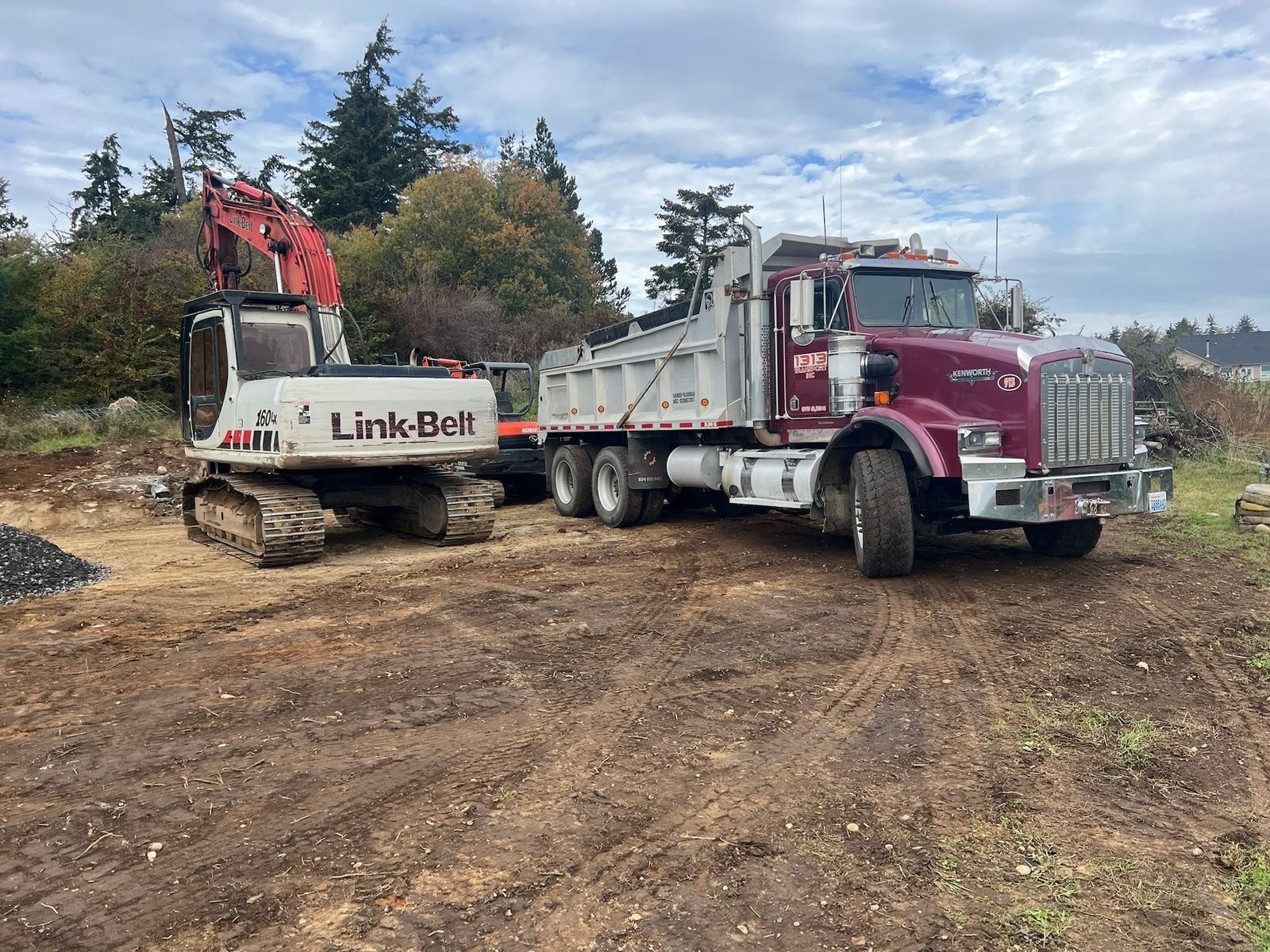 A white Link-Belt excavator parked next to a maroon and white dump truck on a dirt construction site.