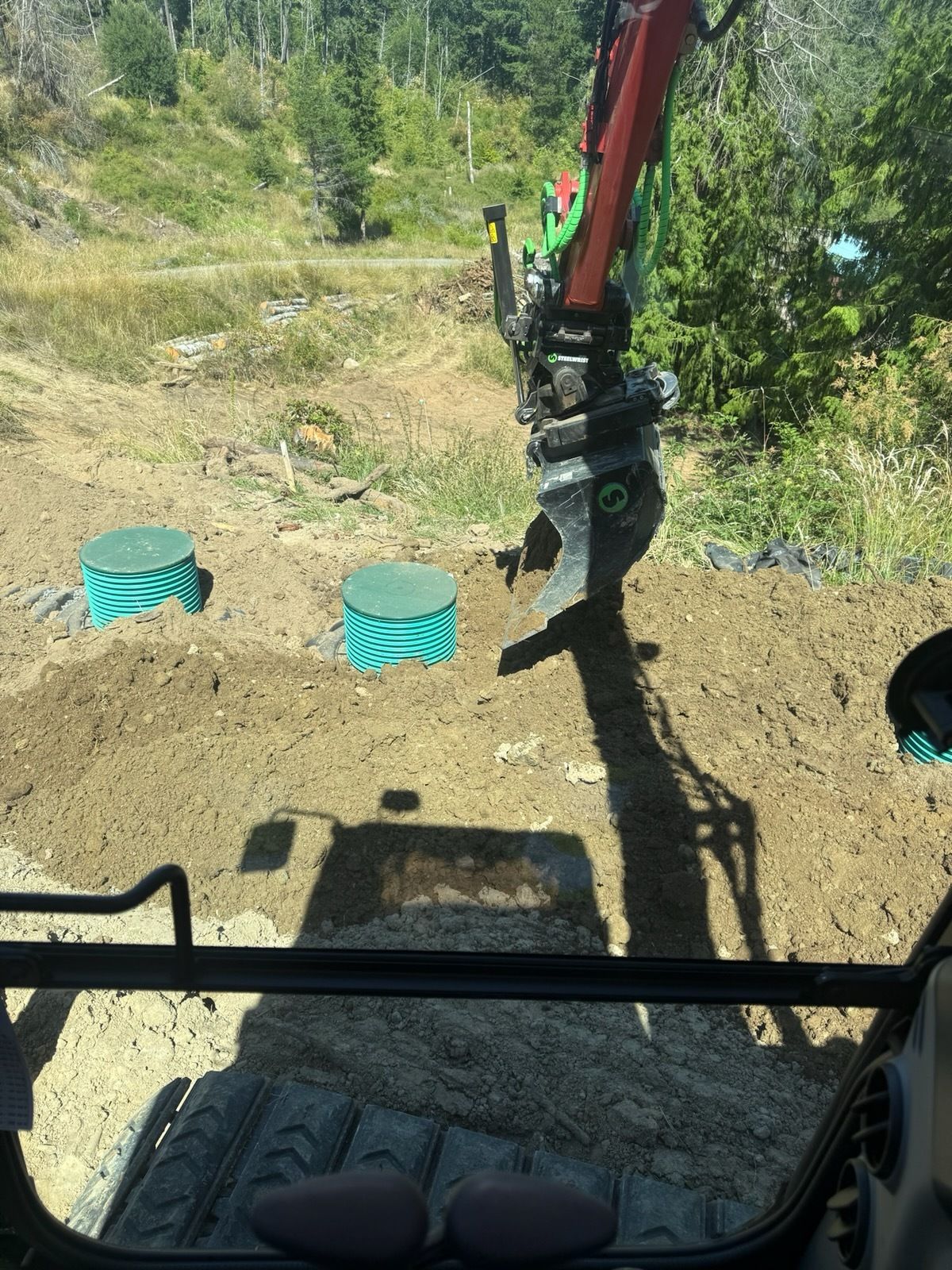View from an excavator cab showing a mechanical arm working near two green, circular septic tank lids in a dirt field.