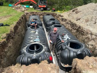 View from an excavator cab showing a mechanical arm working near two green, circular septic tank lids in a dirt field.