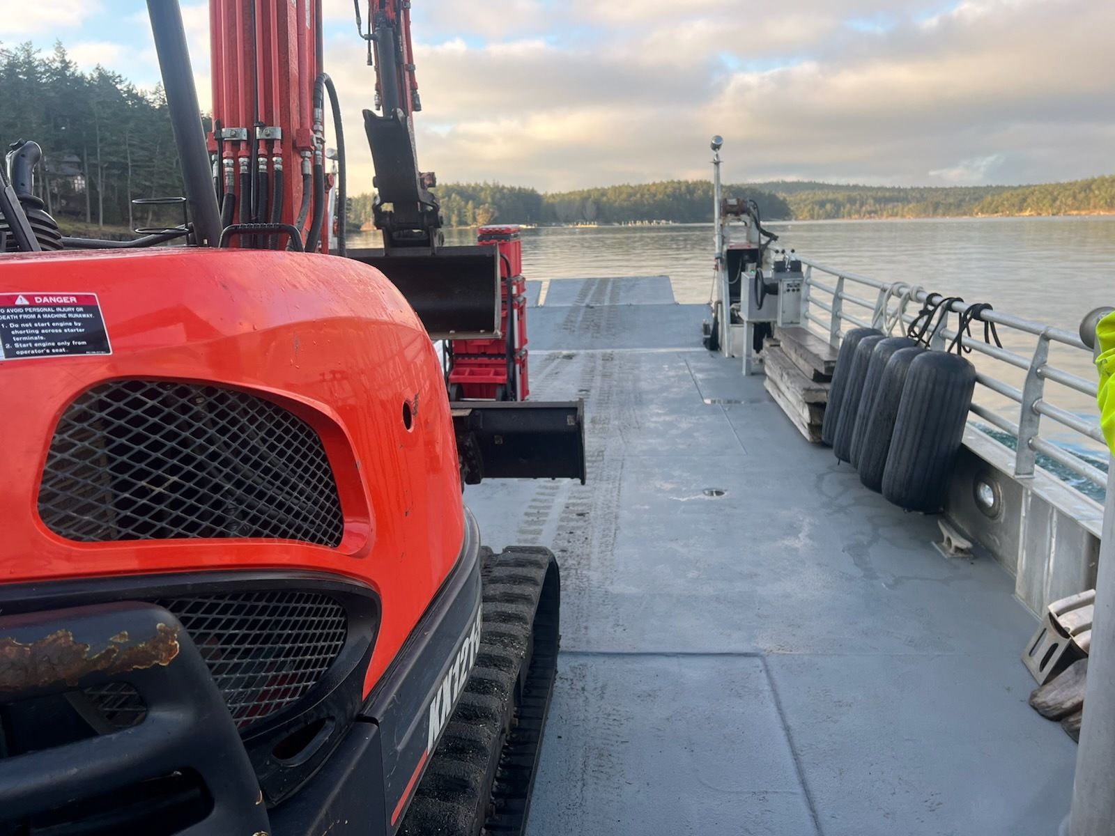A bright orange excavator sits on the deck of a ferry crossing calm water toward a tree-lined shore.