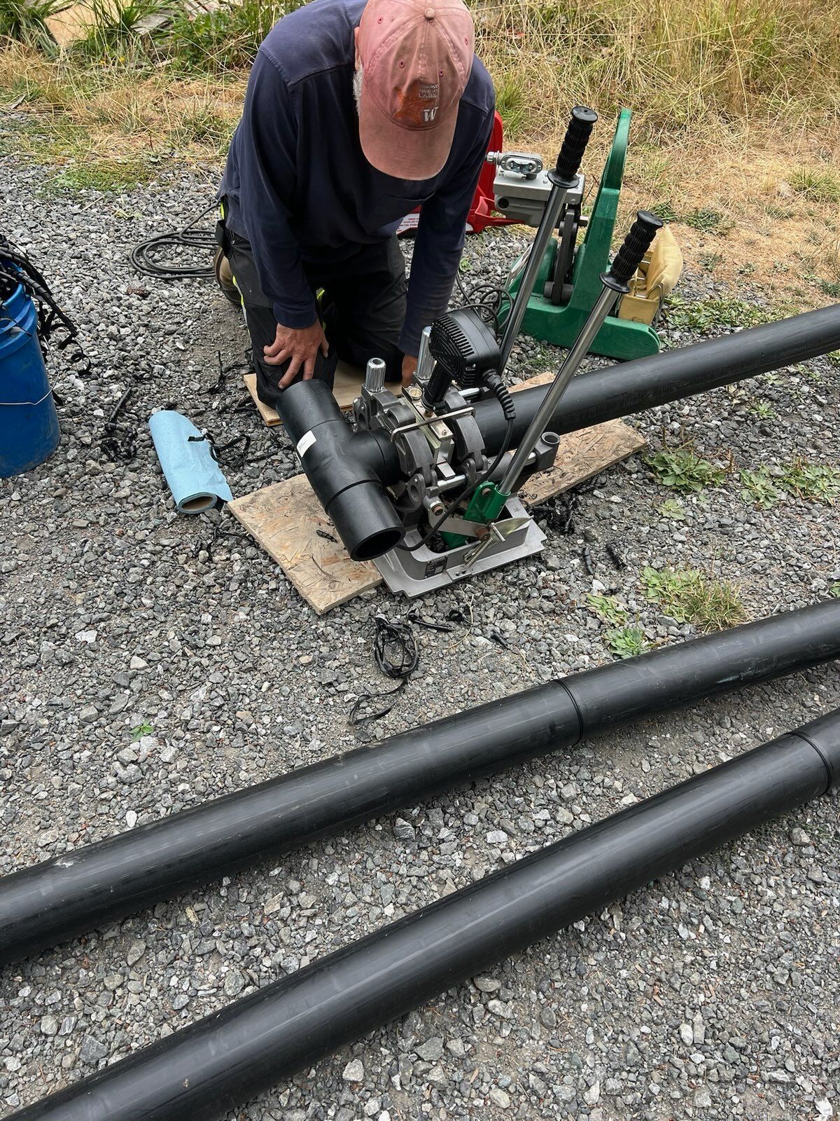A person wearing a hat kneeling on gravel, using a machine to join black HDPE pipe sections.
