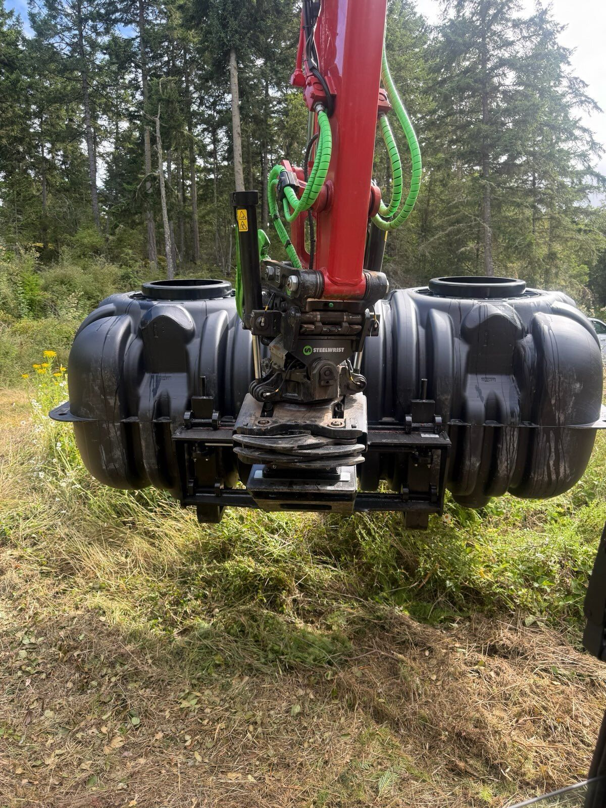 A red excavator arm holds a black plastic septic tank over a grassy, wooded construction site.