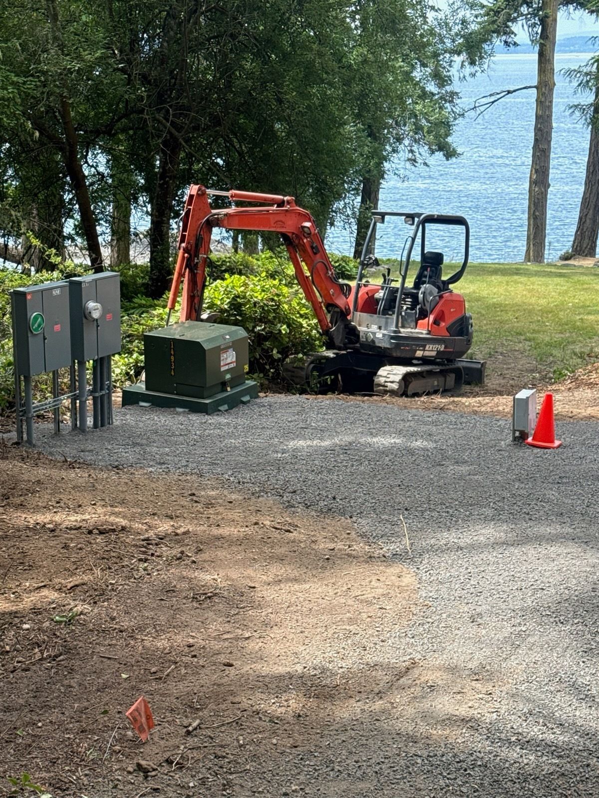 An orange mini excavator parked on a gravel patch near electrical utility boxes and a view of the water.