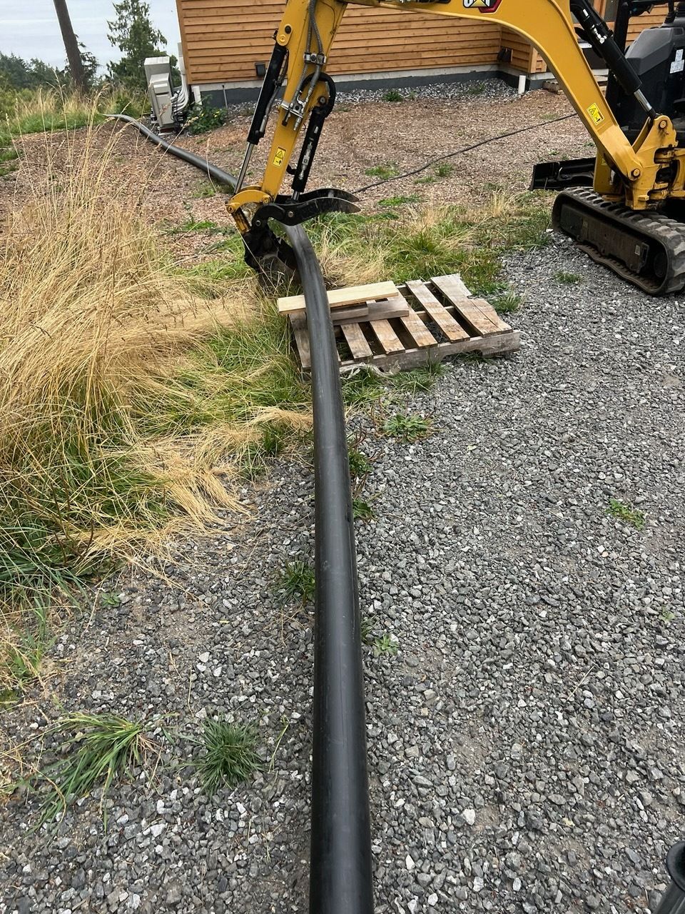 A yellow excavator gripping a long, black pipe over a wooden pallet on a gravel surface next to a building.