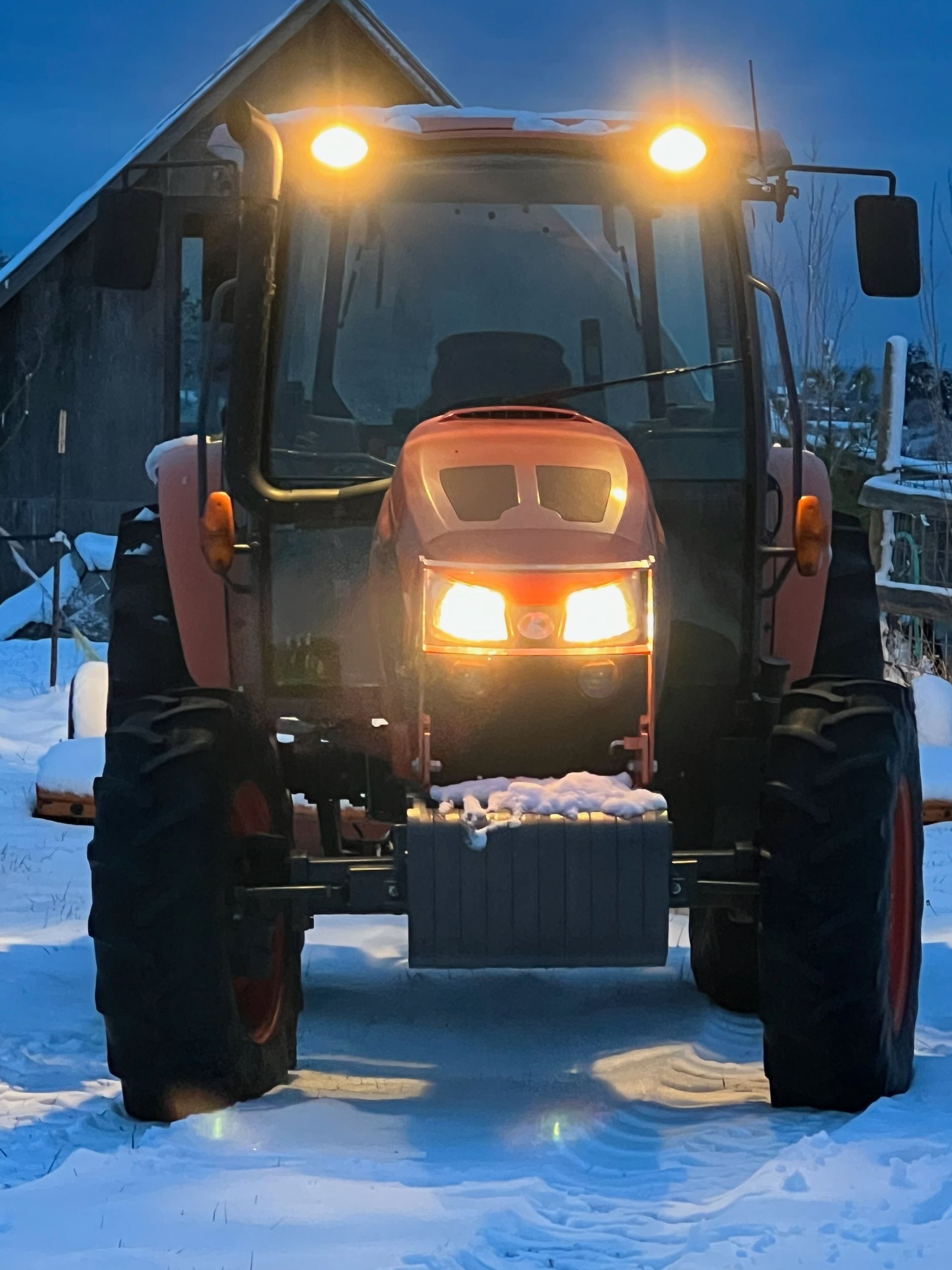 A front view of an orange Kubota tractor with its headlights on, parked in a snow-covered field during twilight.