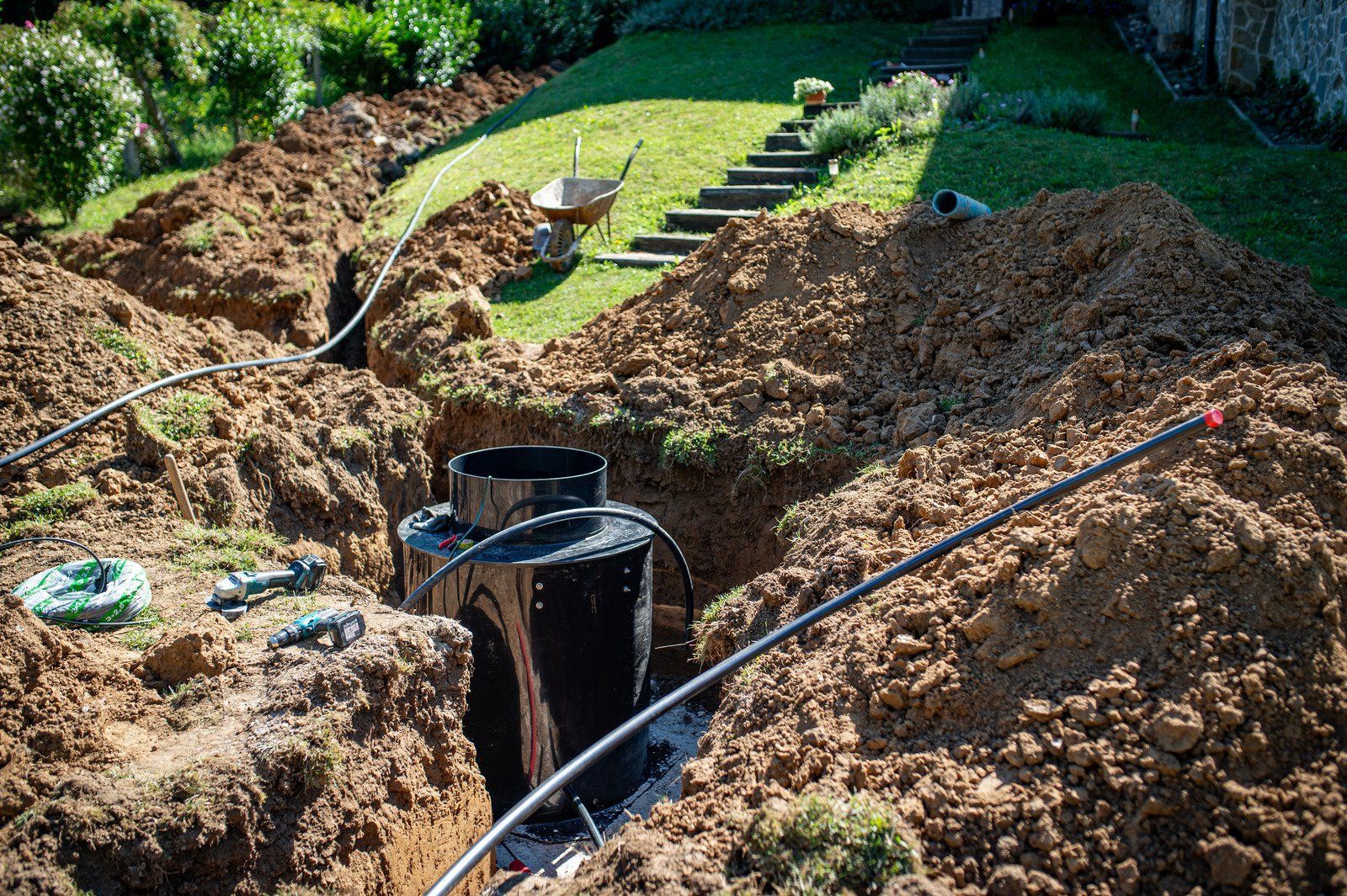 A large black tank is installed in a deep trench dug into a green lawn, surrounded by piles of excavated earth and piping.