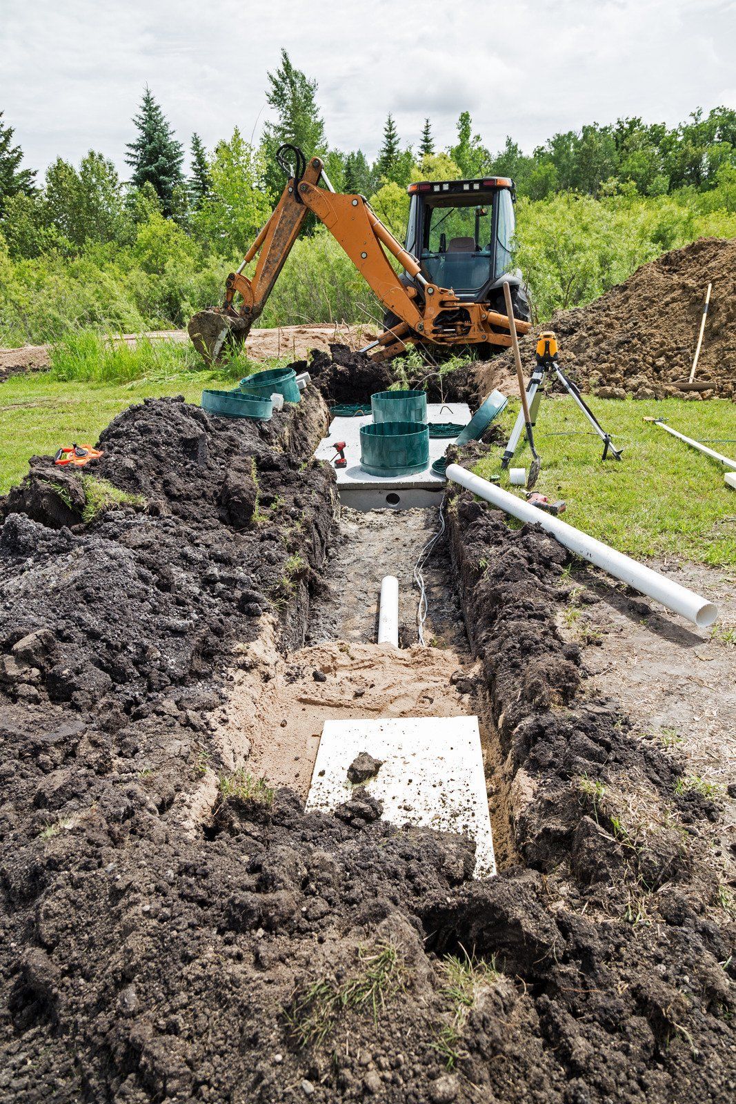 An orange backhoe parked near a long excavation trench with a concrete septic tank and white pipes in a grassy field.