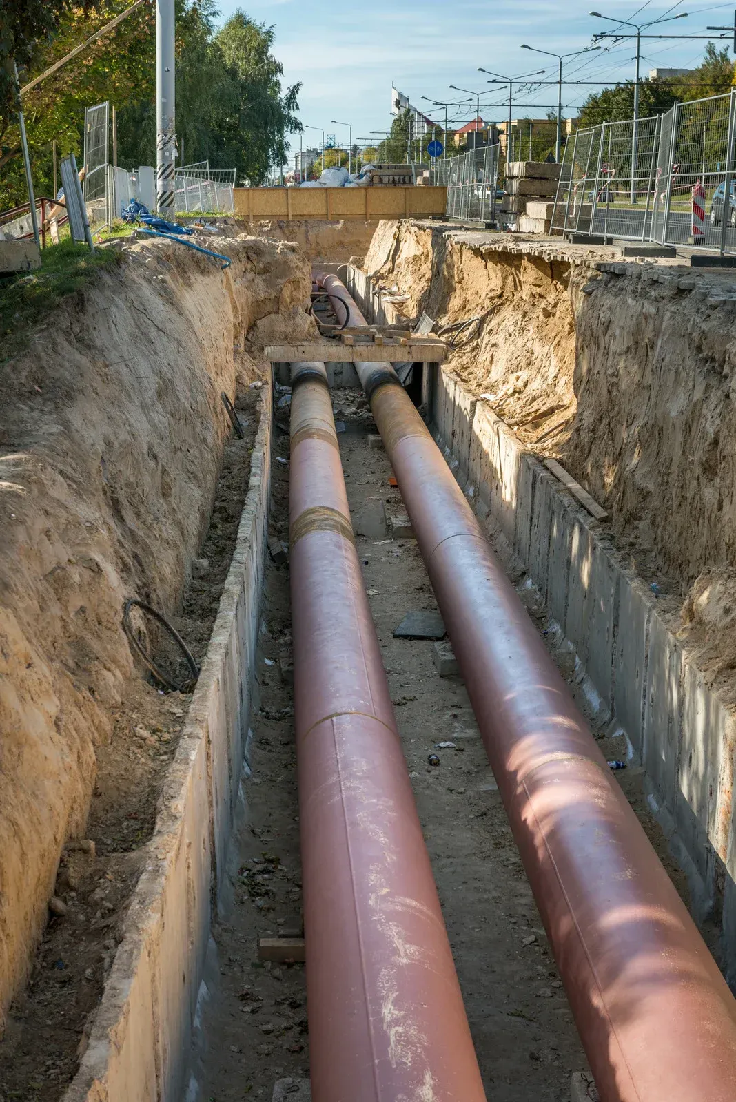 Two large, red-brown pipes run parallel through a deep, open dirt trench at a construction site.