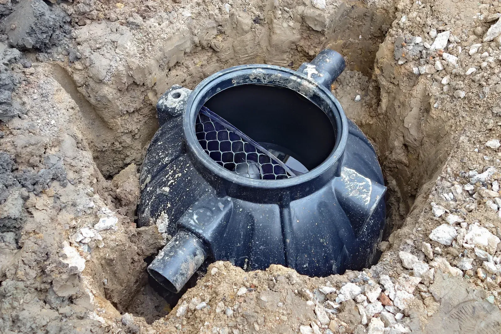 A black plastic septic or drainage basin installed in a dug earth pit, featuring a visible interior mesh filter screen.