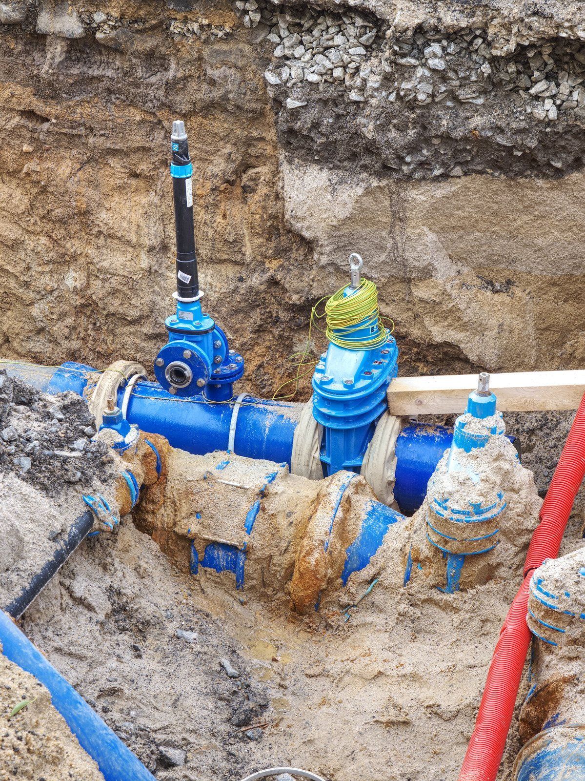 Blue water pipes and valves with an upright control handle partially buried in an excavated construction trench.