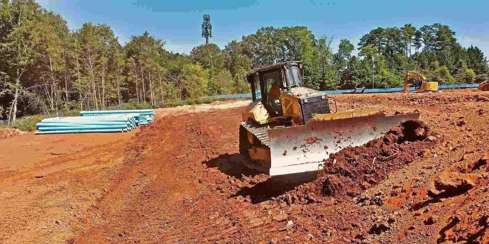 A yellow bulldozer grades red dirt on a construction site with blue pipes stacked in the background.