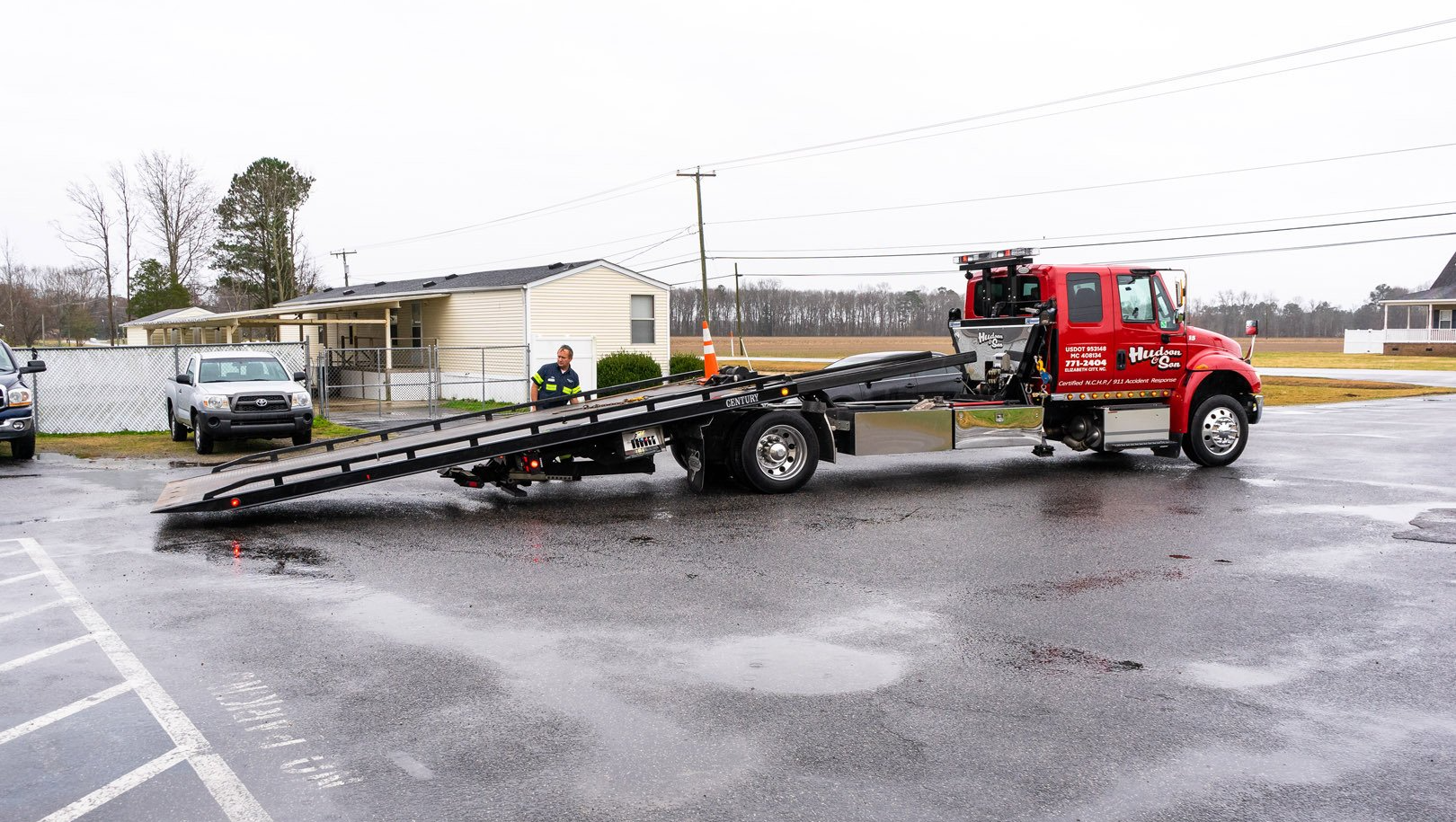 Tow truck extending its flatbed in a wet parking lot next to a small building and a pickup truck.