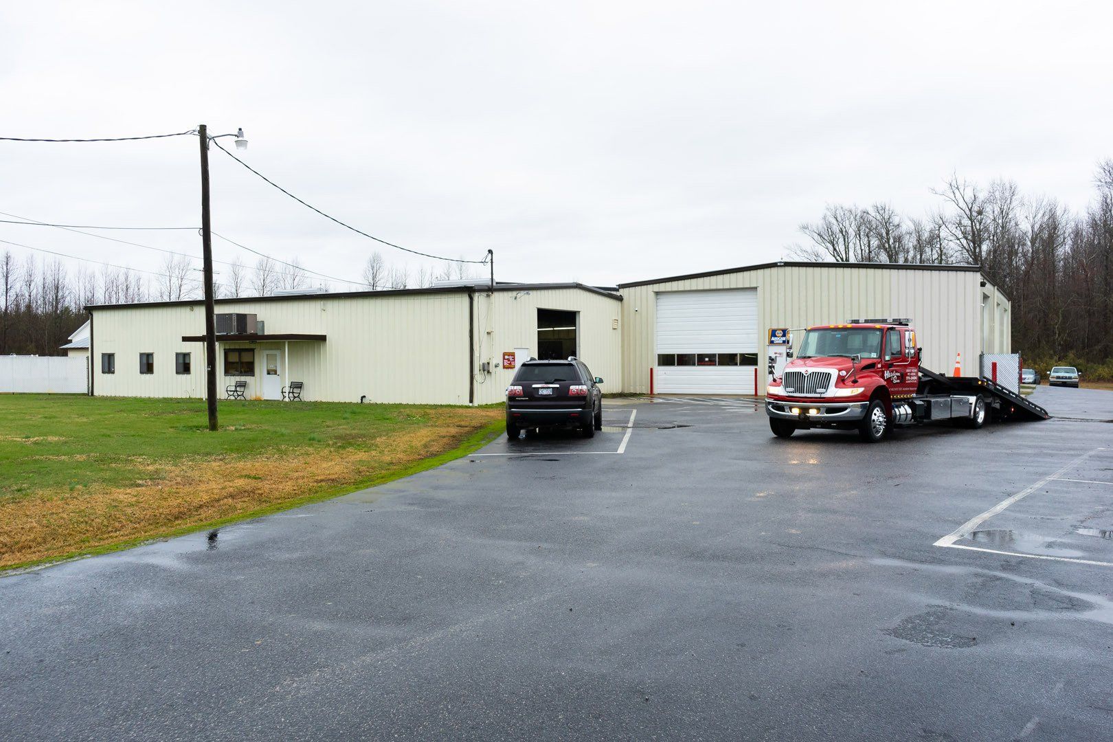 A tow truck prepares to load a dark SUV in front of a tan metal building with an open garage door; overcast day.