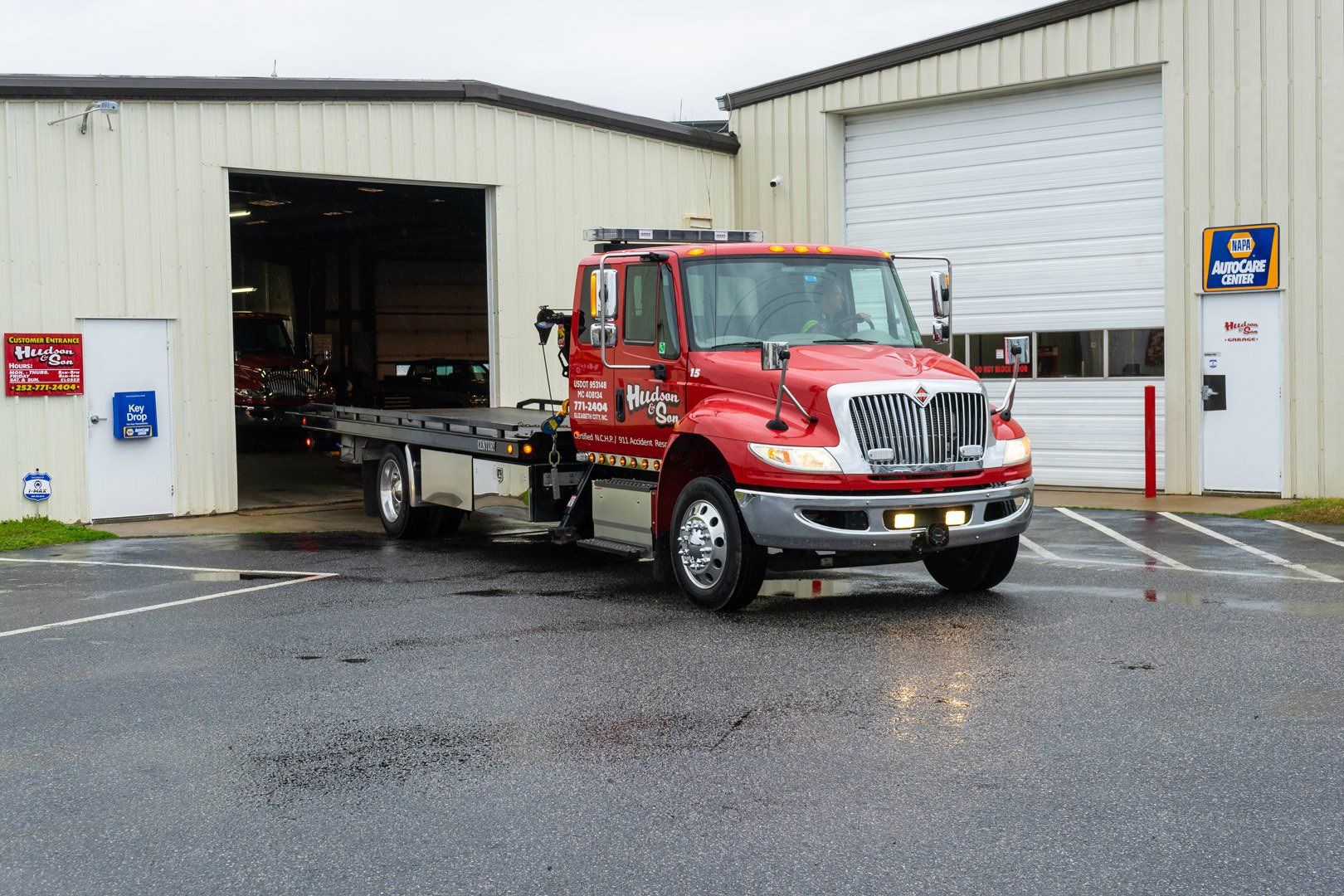 Red tow truck outside a repair shop bay on a wet, paved surface.