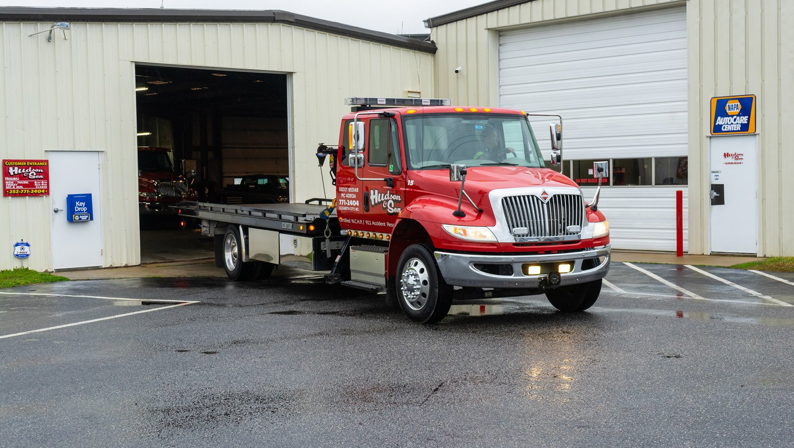 Red tow truck parked in front of a garage, doors open. Wet pavement.