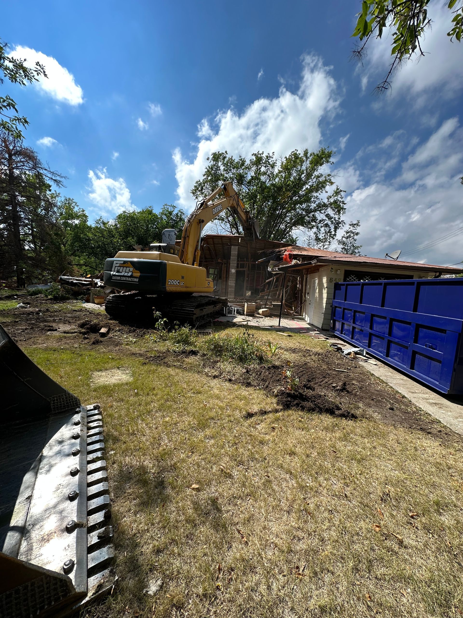 A yellow excavator is demolishing a house in a yard.