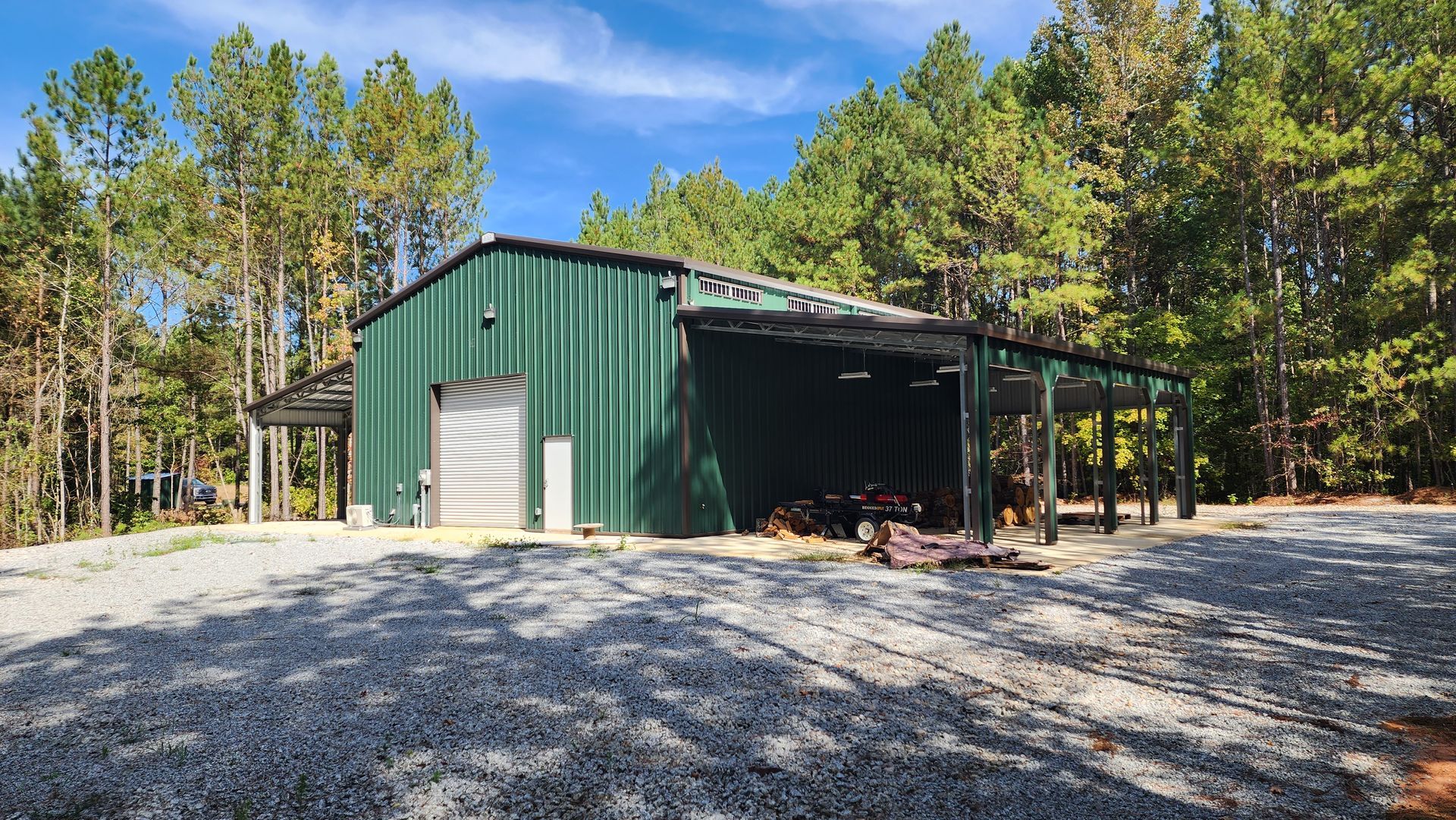 A large metal garage is sitting on top of a gravel driveway in the middle of a forest.