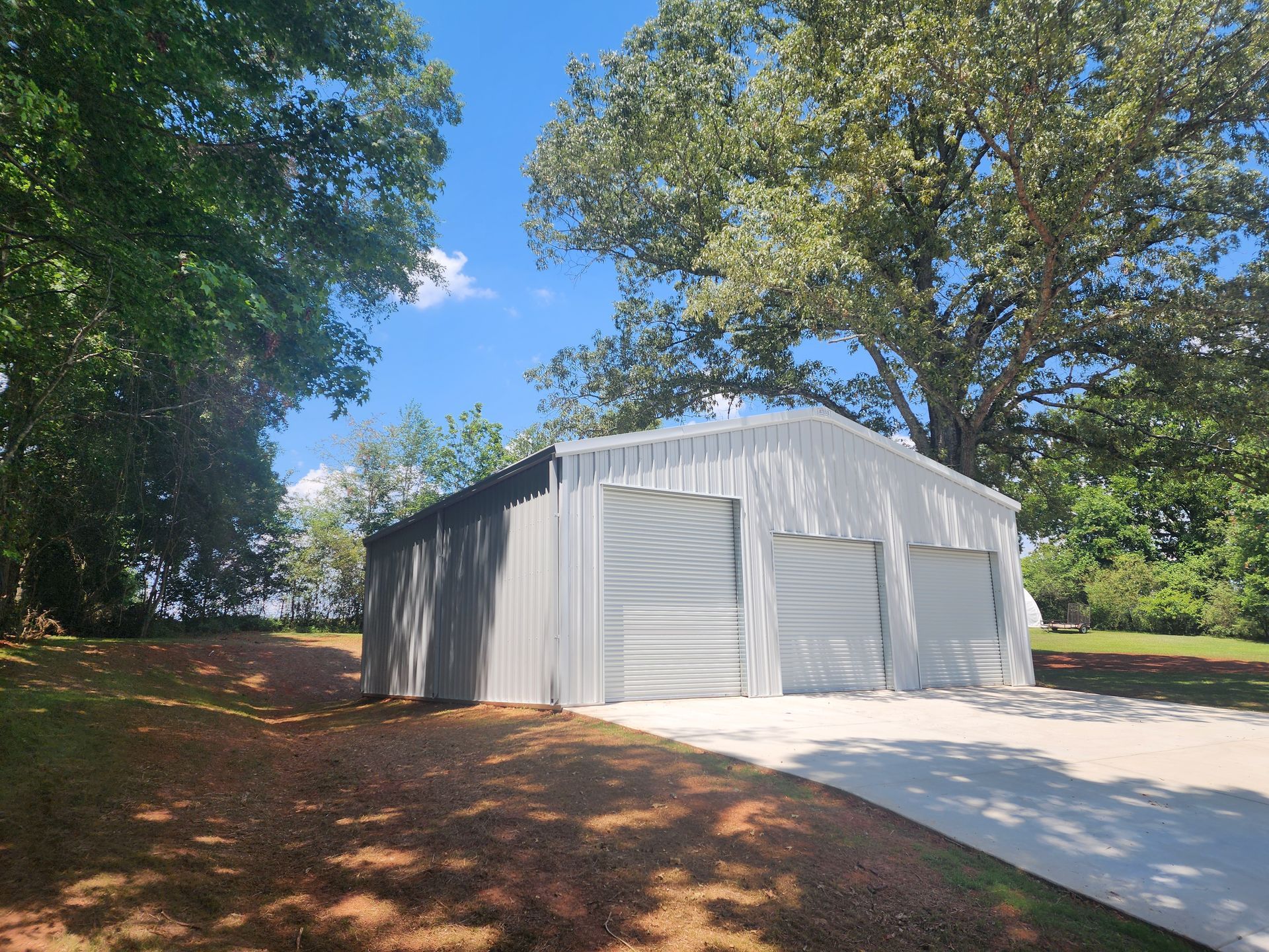 A large metal garage with three garage doors is surrounded by trees and a driveway.