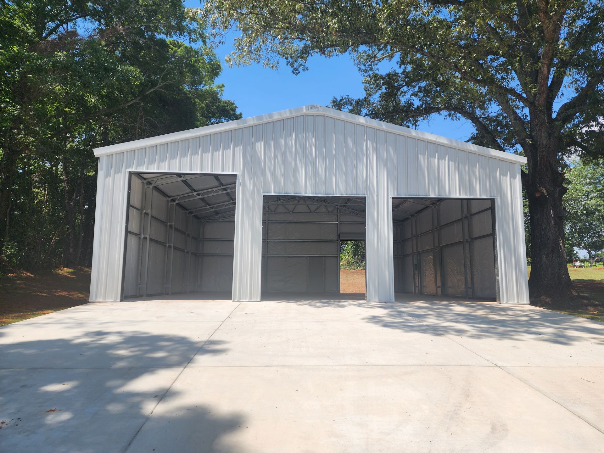 A large white metal building with three garage doors is sitting on top of a concrete driveway.