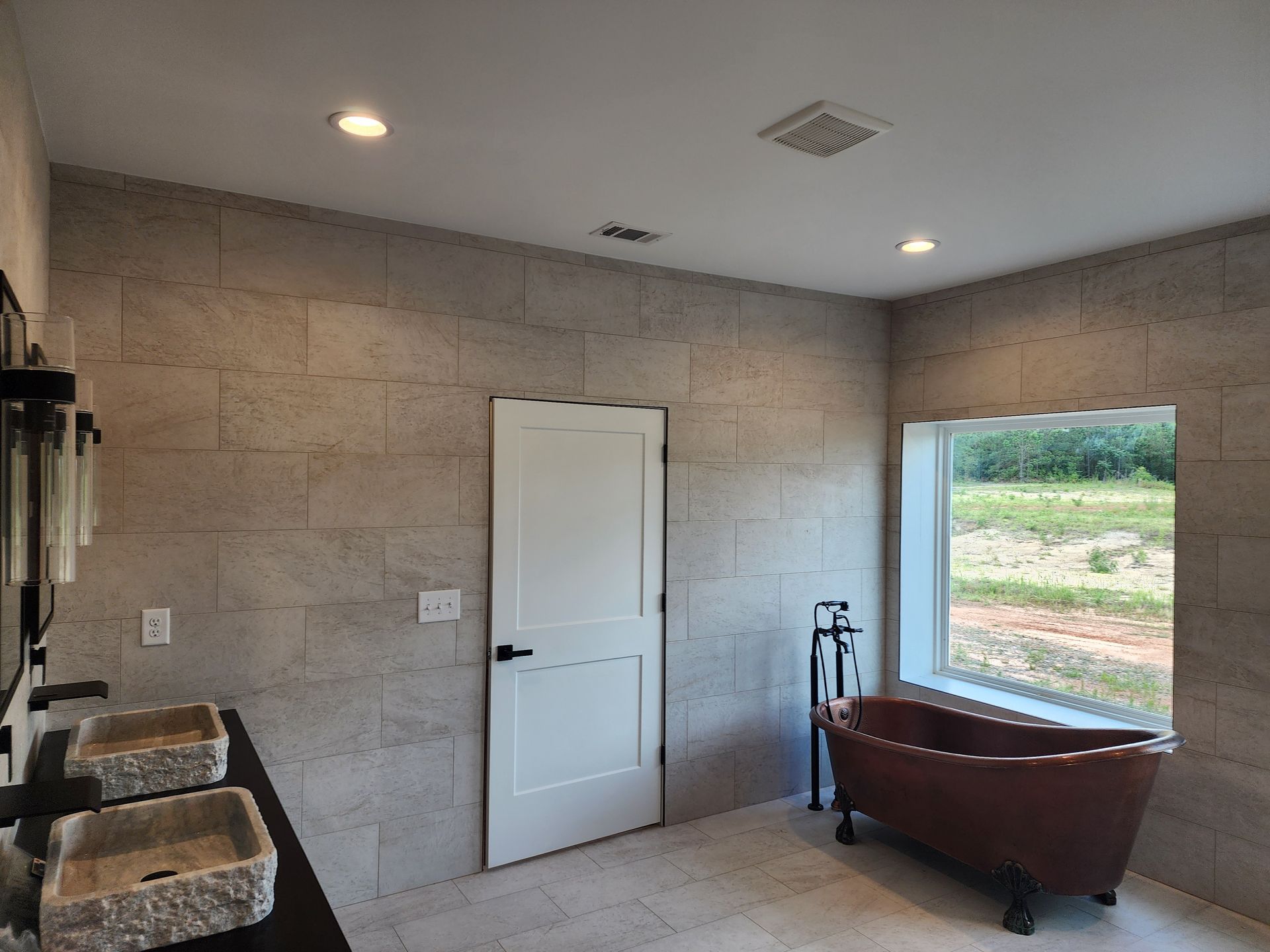 Modern bathroom with stone tile walls, white door, copper bathtub, and window with outdoor view.