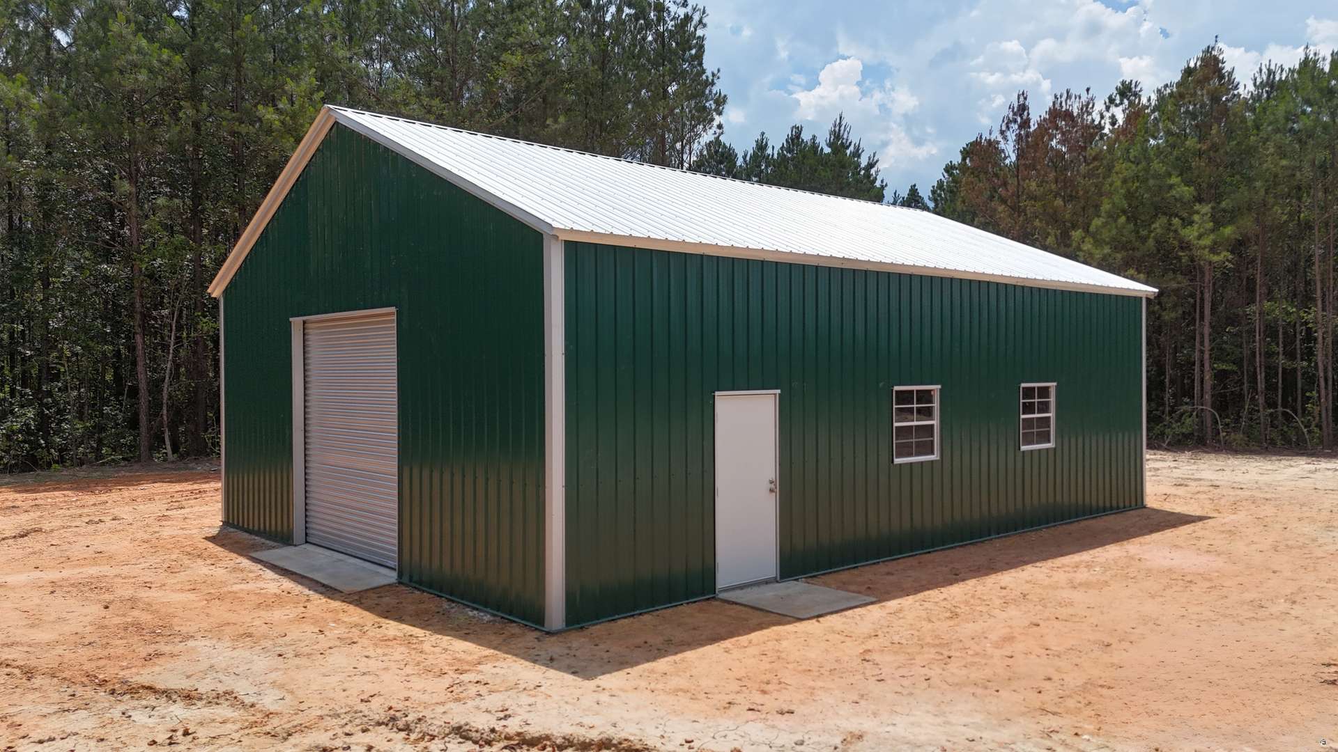 Green metal barn-like building with white roof and doors on a dirt lot, surrounded by trees.