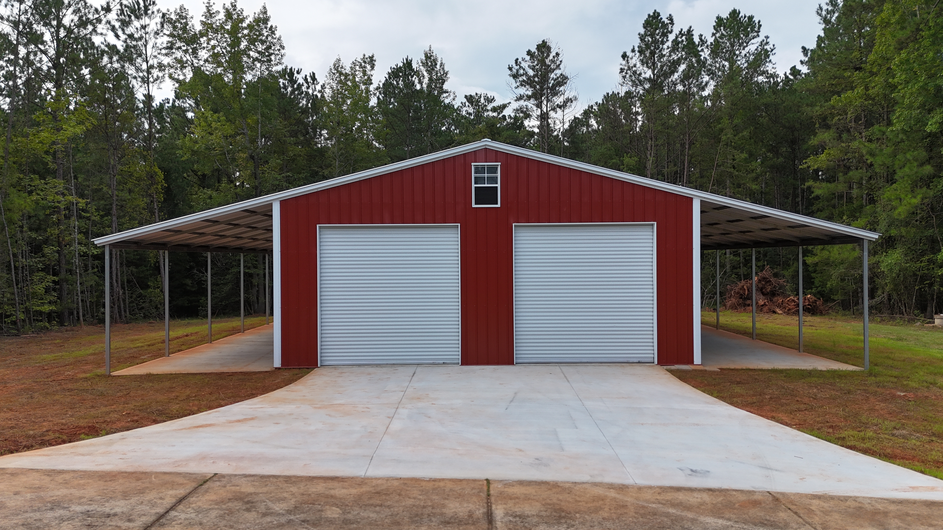 Red metal garage with two white doors and side carports on a concrete driveway, surrounded by trees.