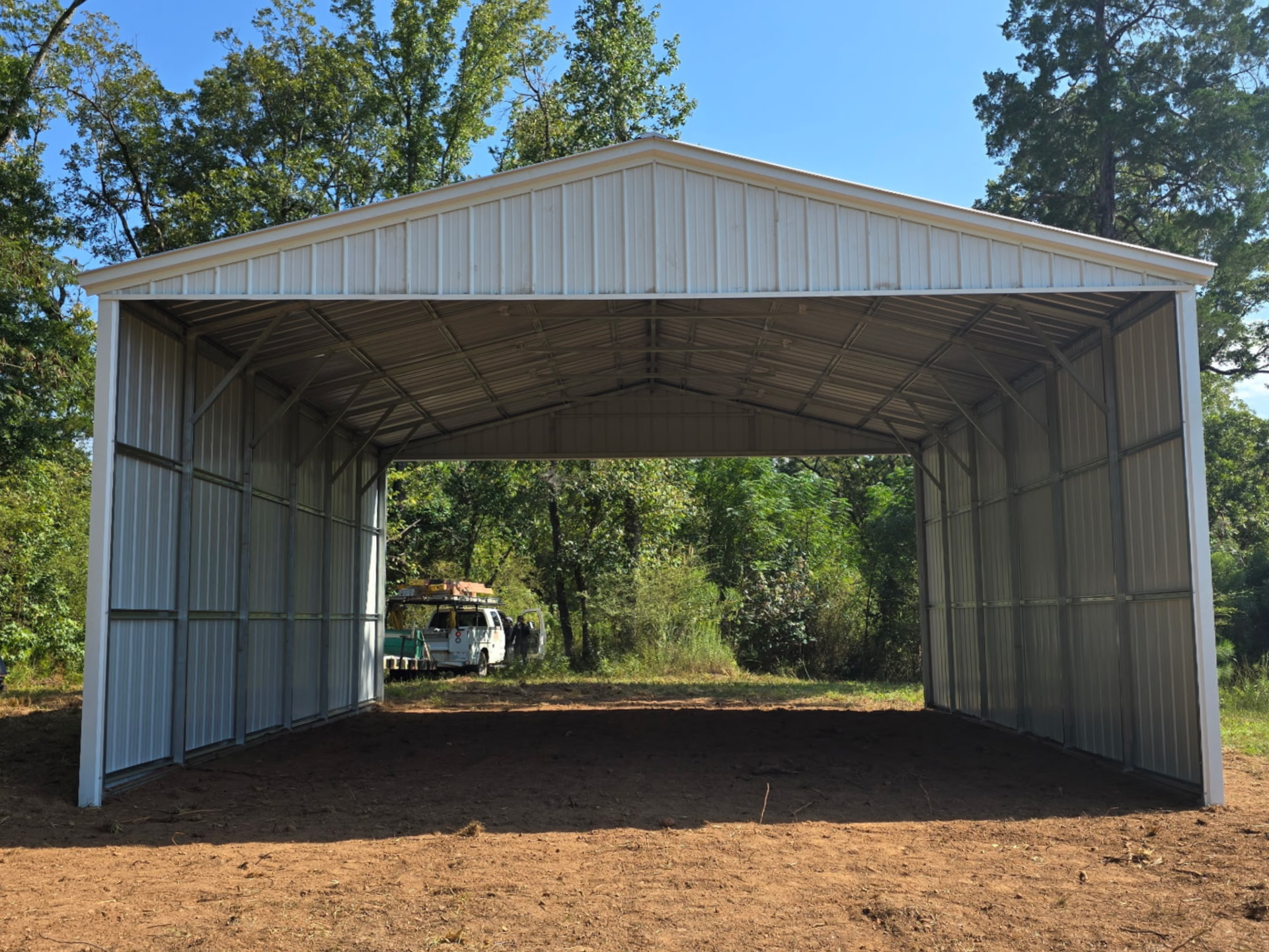 Open-sided white metal barn or carport on a dirt lot, with trees in the background.