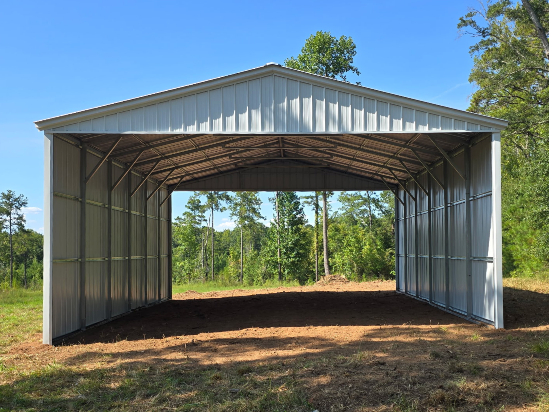 Open-sided metal shed with a white roof and dirt floor, set among trees under a blue sky