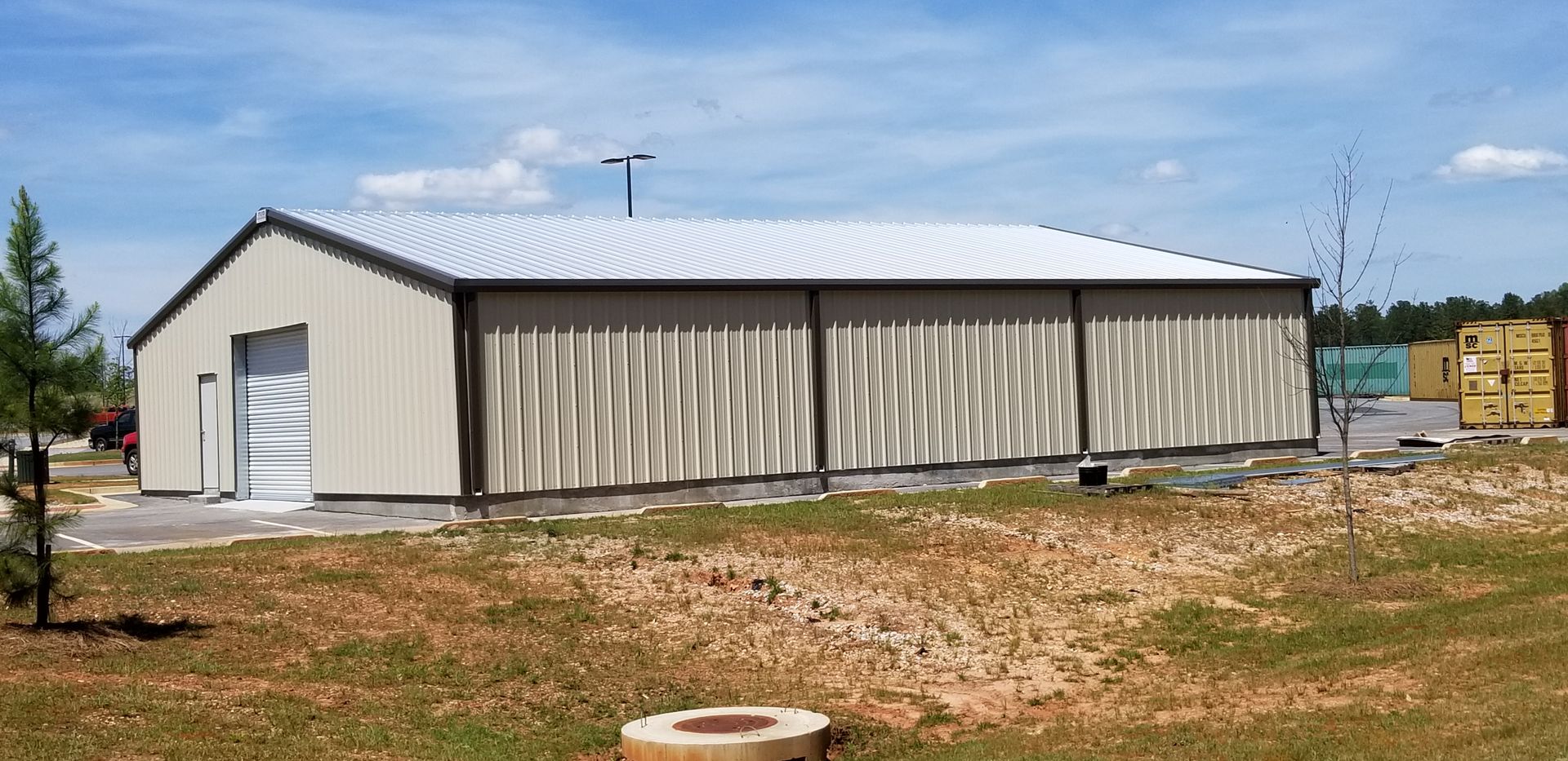 A metal industrial building with a light tan exterior and corrugated silver roof. Blue sky and trees in the background.