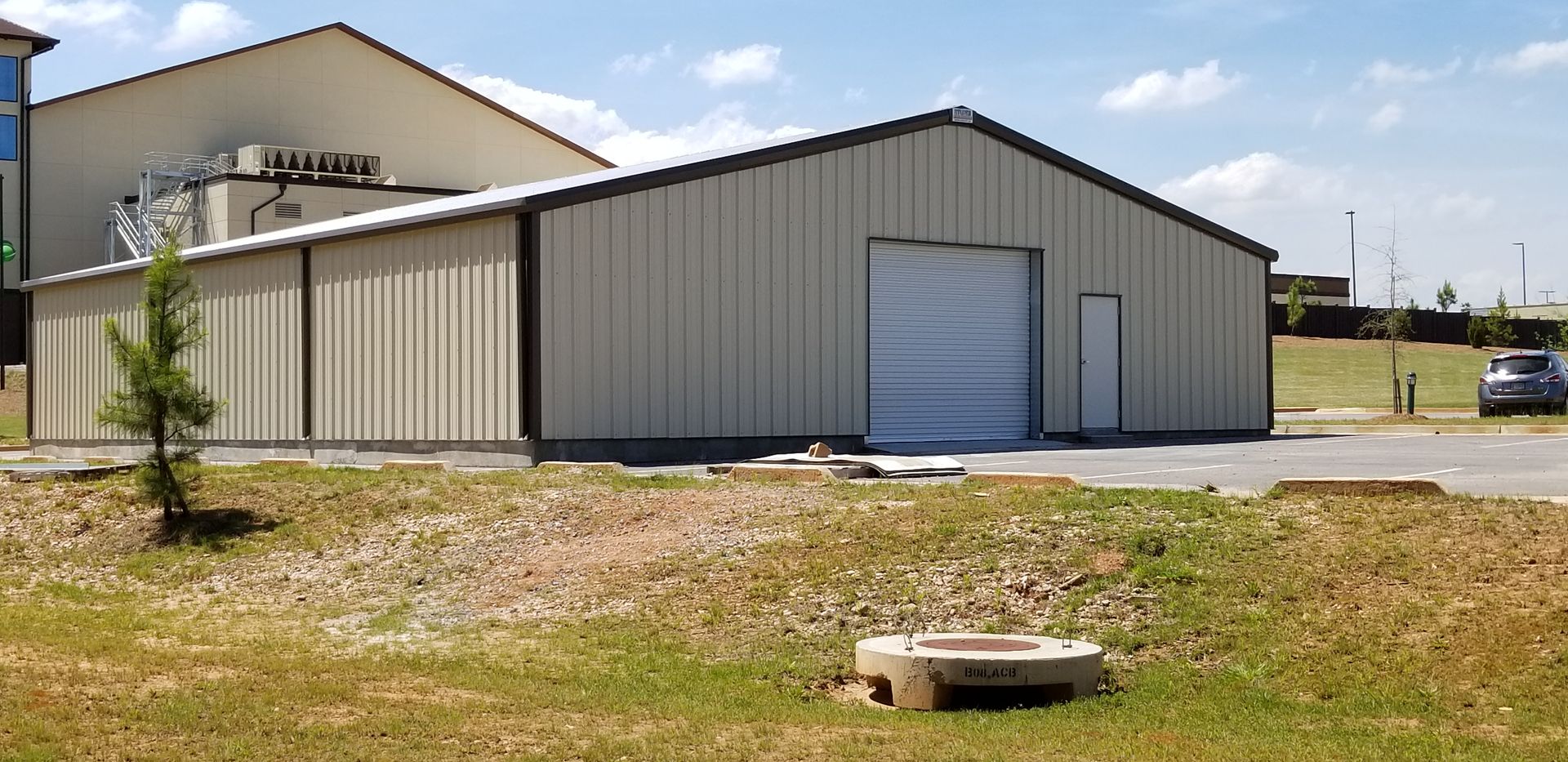 A beige metal building with a closed garage door sits on a grassy area under a blue sky.