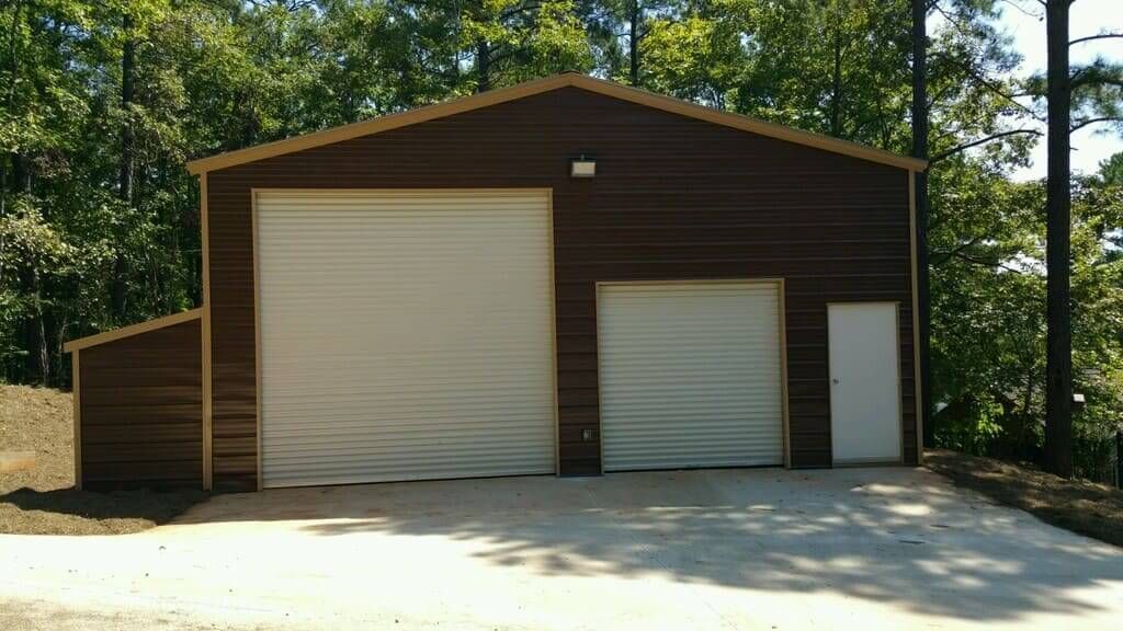 A brown garage with two white garage doors is sitting in the middle of a forest.