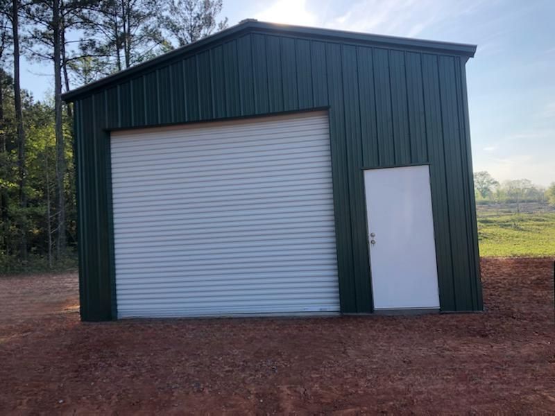 A green metal garage with a white door is sitting in the middle of a dirt field.