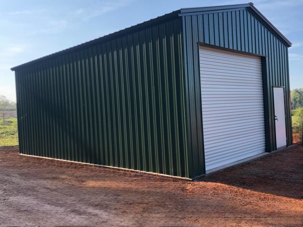 A green metal building with a white garage door is sitting on top of a dirt field.