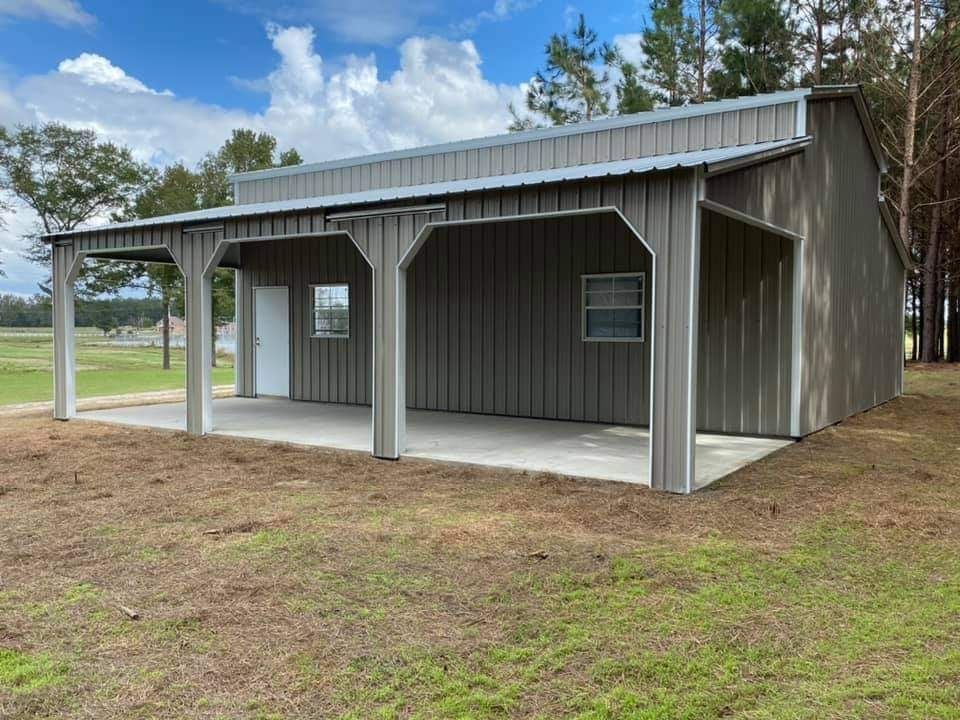 A gray metal building with a porch and a driveway is sitting in the middle of a grassy field.
