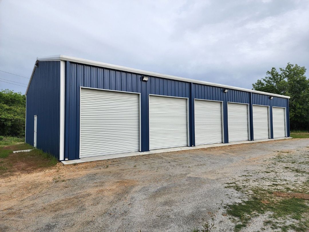 A blue metal building with white garage doors is sitting on top of a gravel road.