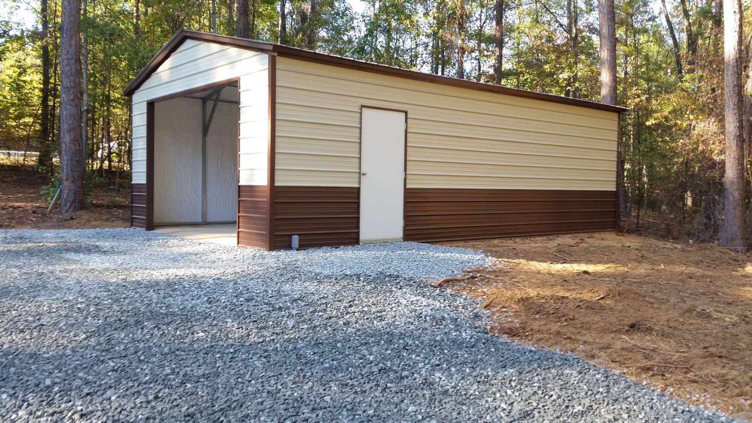 A metal garage is sitting on top of a gravel driveway in the woods.