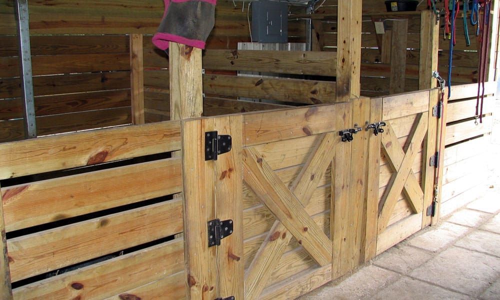 A horse is standing in a wooden stable behind a wooden gate.