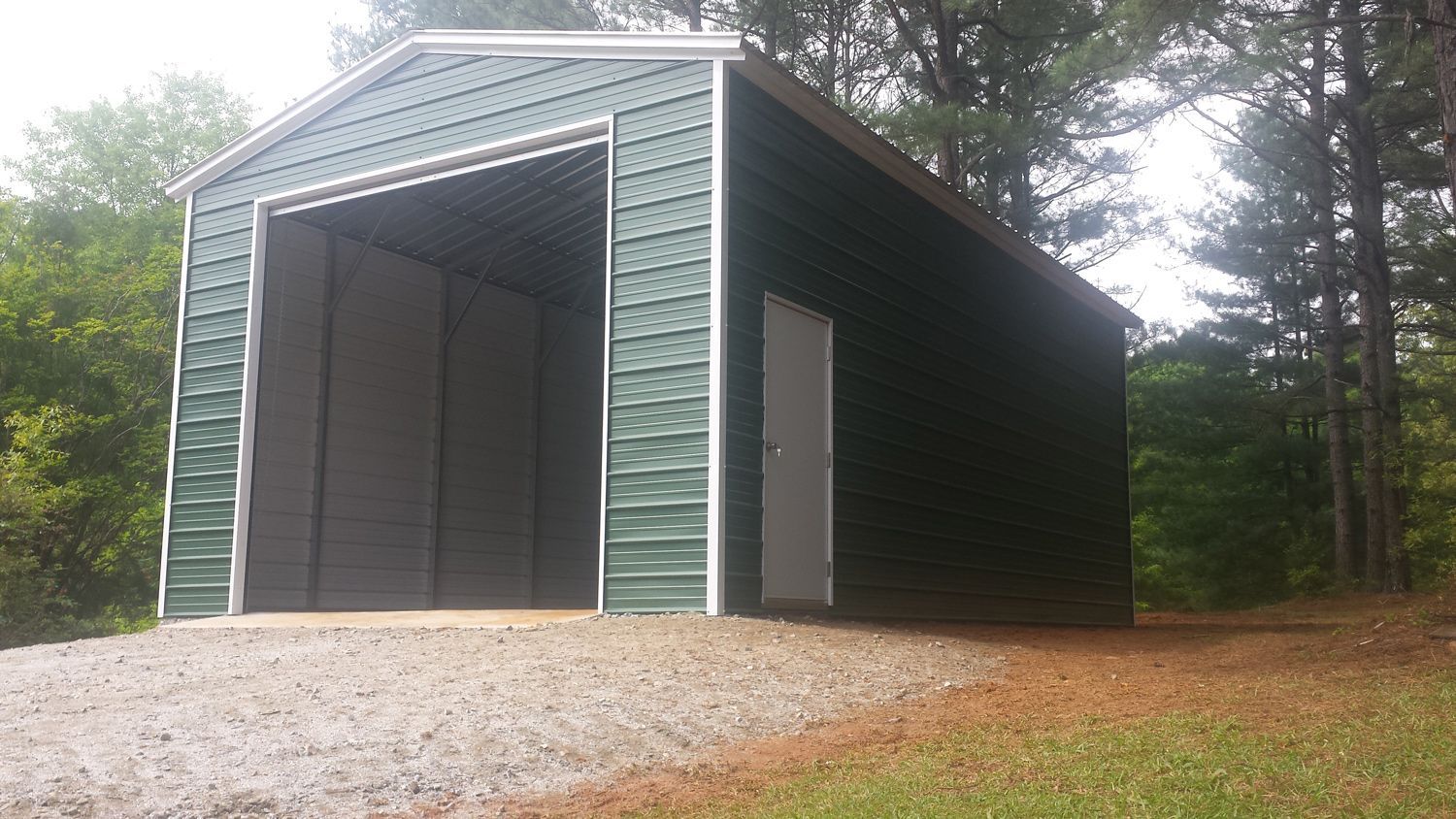 A green metal garage with a large door is sitting on top of a gravel driveway.