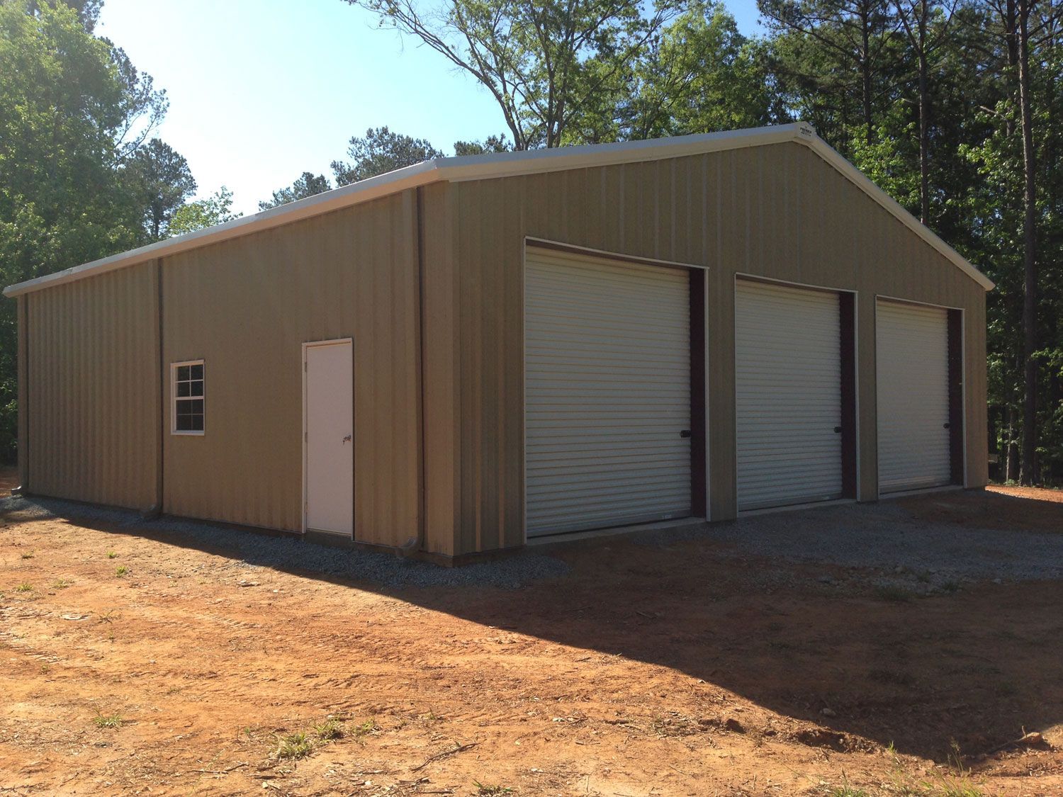 A large metal building with three garage doors is sitting in the middle of a dirt field.