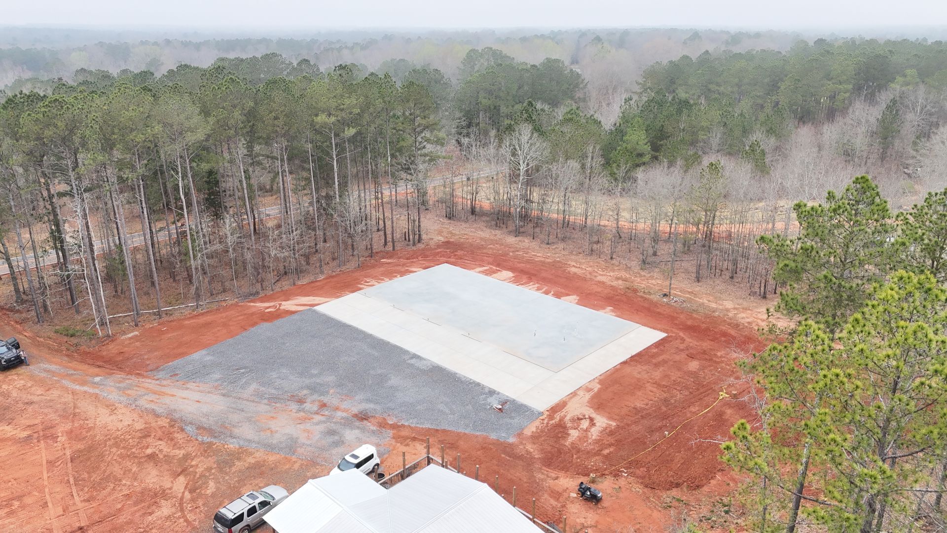 Aerial view of a red dirt construction site with a large gray concrete slab beside a pine forest.