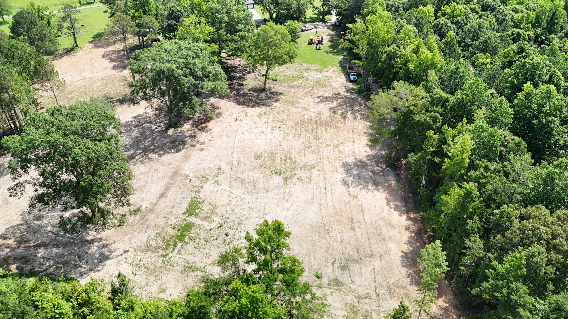 Aerial view of a cleared dirt field bordered by dense green trees and scattered shade trees.