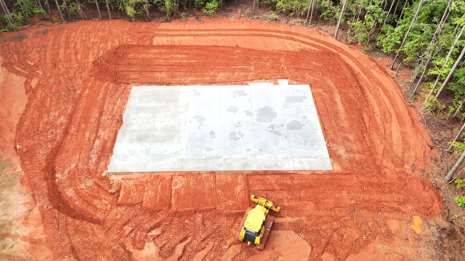 Aerial view of a dirt construction site with a large concrete slab and a small yellow machine nearby.