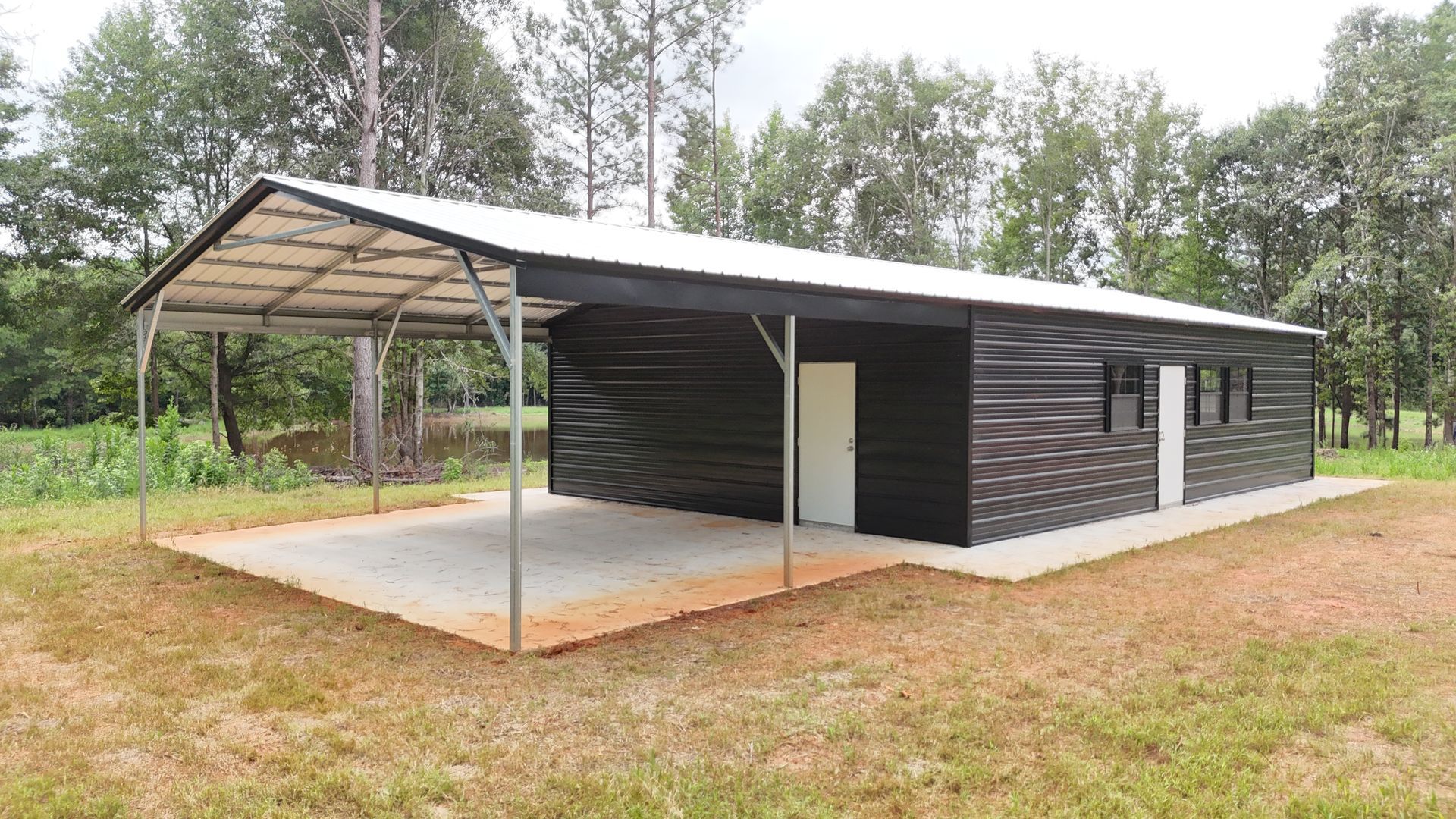 A black metal building with a carport, on a concrete pad, surrounded by grass and trees.