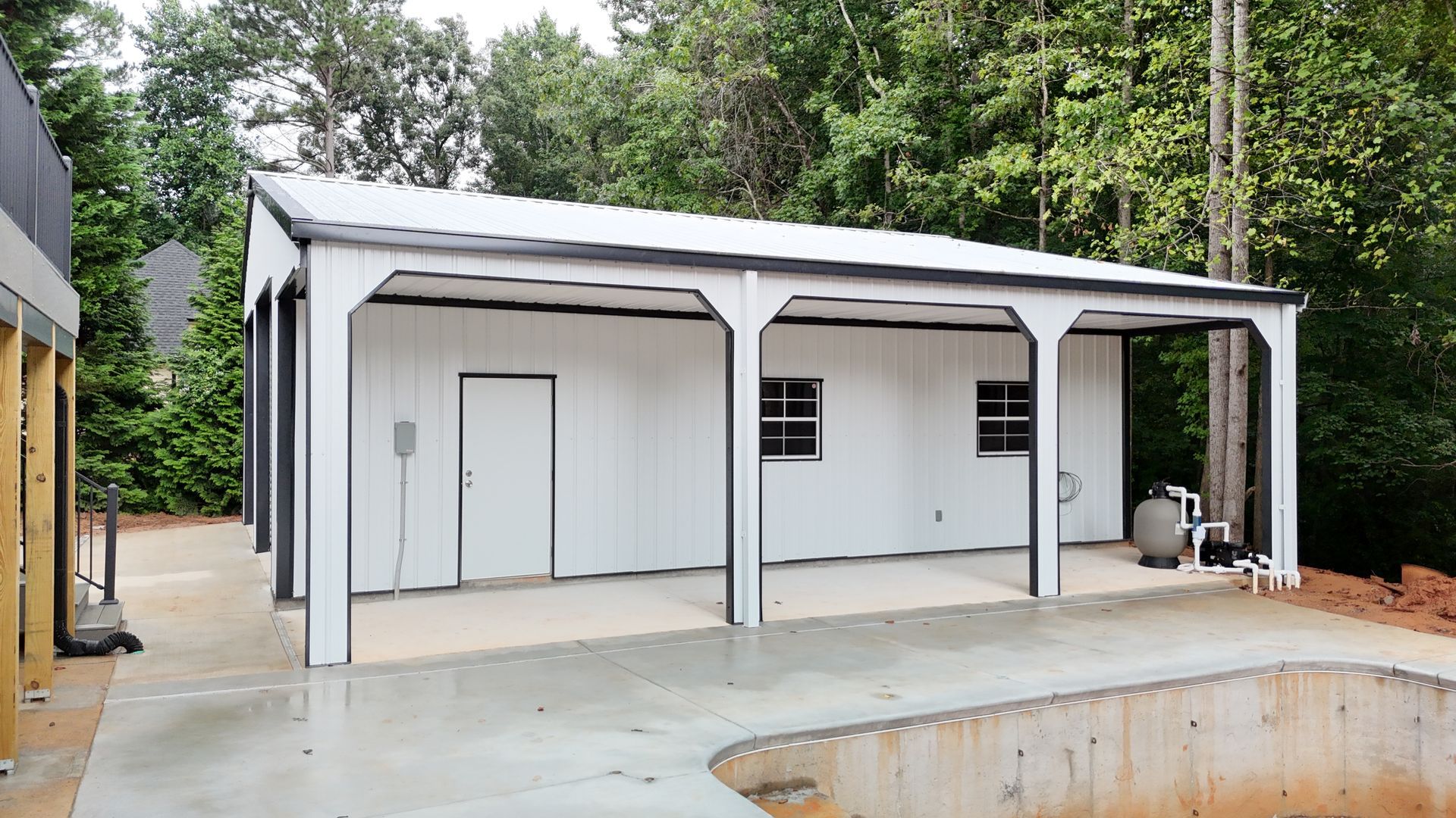 White shed with a gray metal roof and black trim. Features an attached covered patio. Located near a pool under construction.
