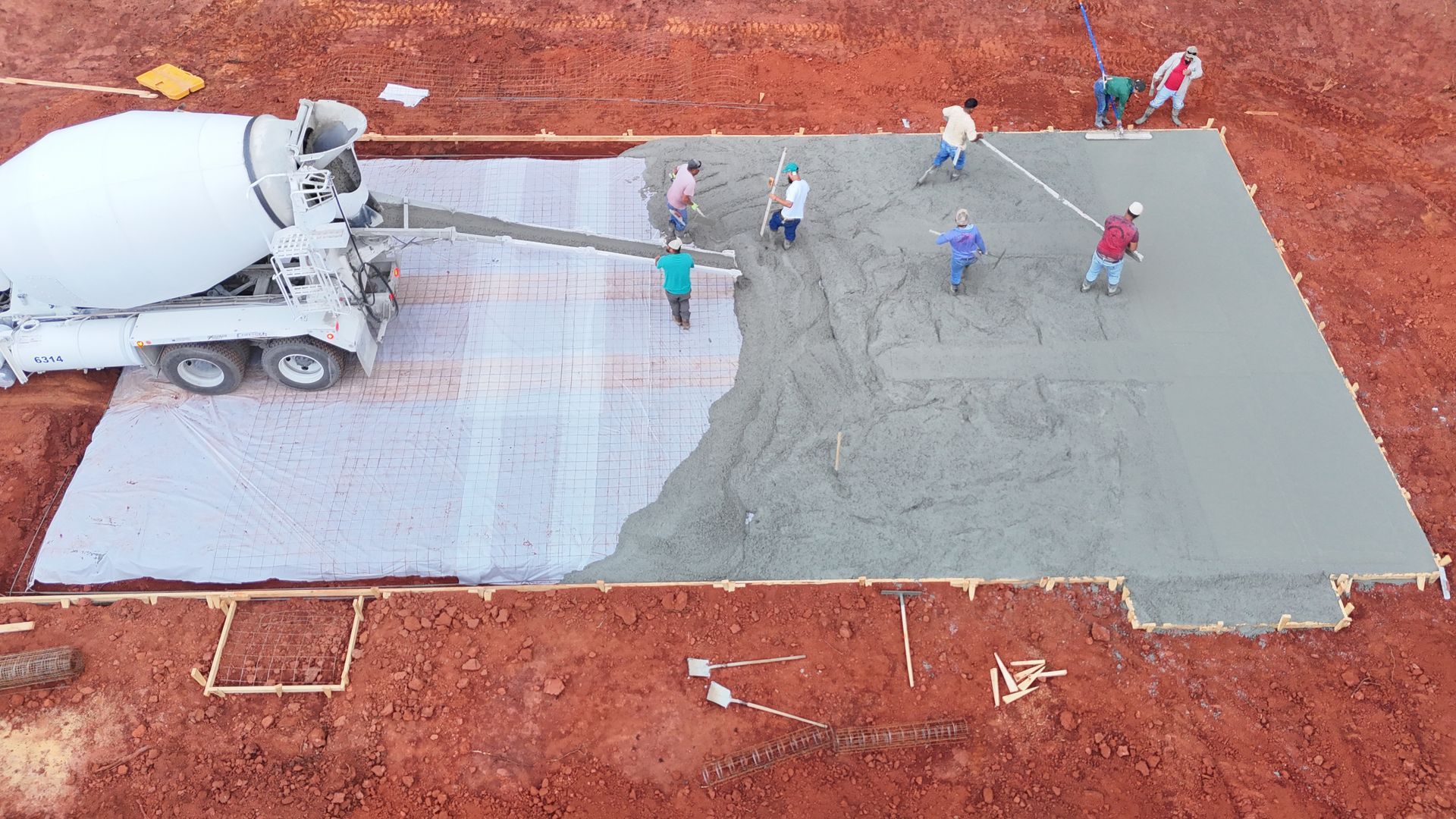 Construction crew pouring concrete for a foundation, using a cement truck and leveling tools, in a dirt setting.