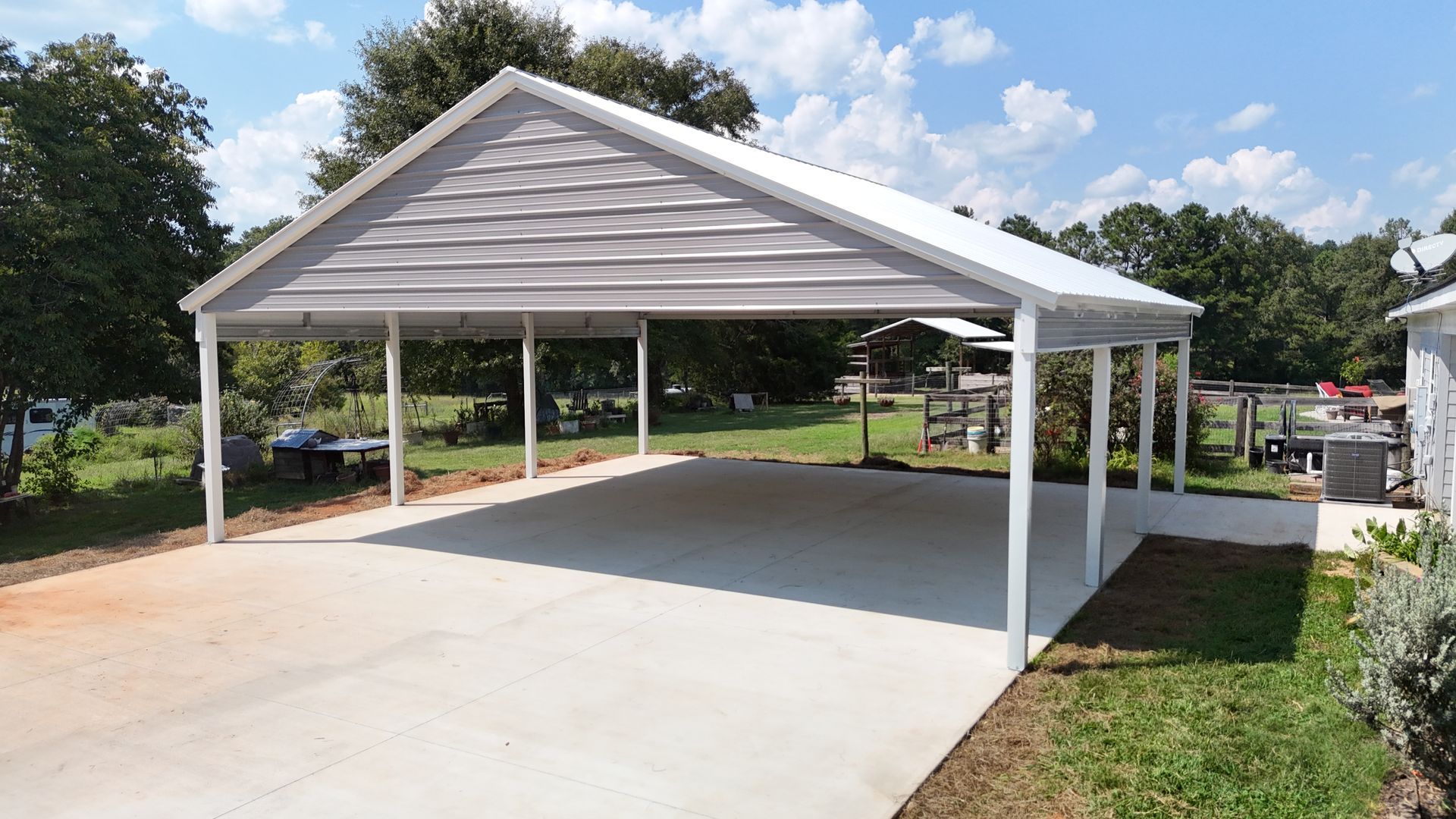 White carport with a gray roof on a concrete pad; green grass and trees in the background.