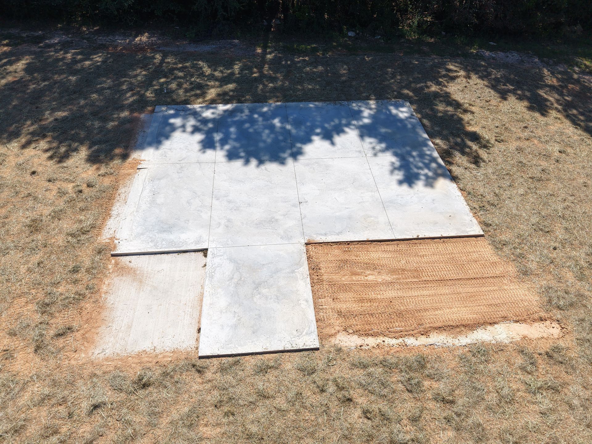 Concrete slabs laid on a dirt lot, forming a T-shaped foundation outline with shadows from trees.